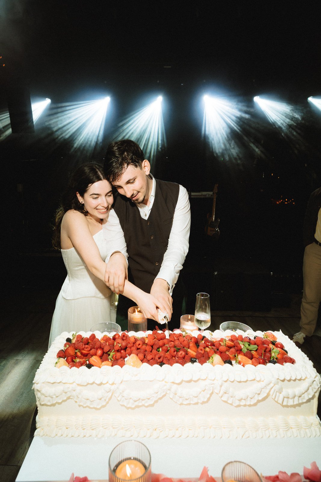 A couple cutting a wedding cake with strawberries and cream, celebrating together in a dimly lit venue with stage lights and candles.