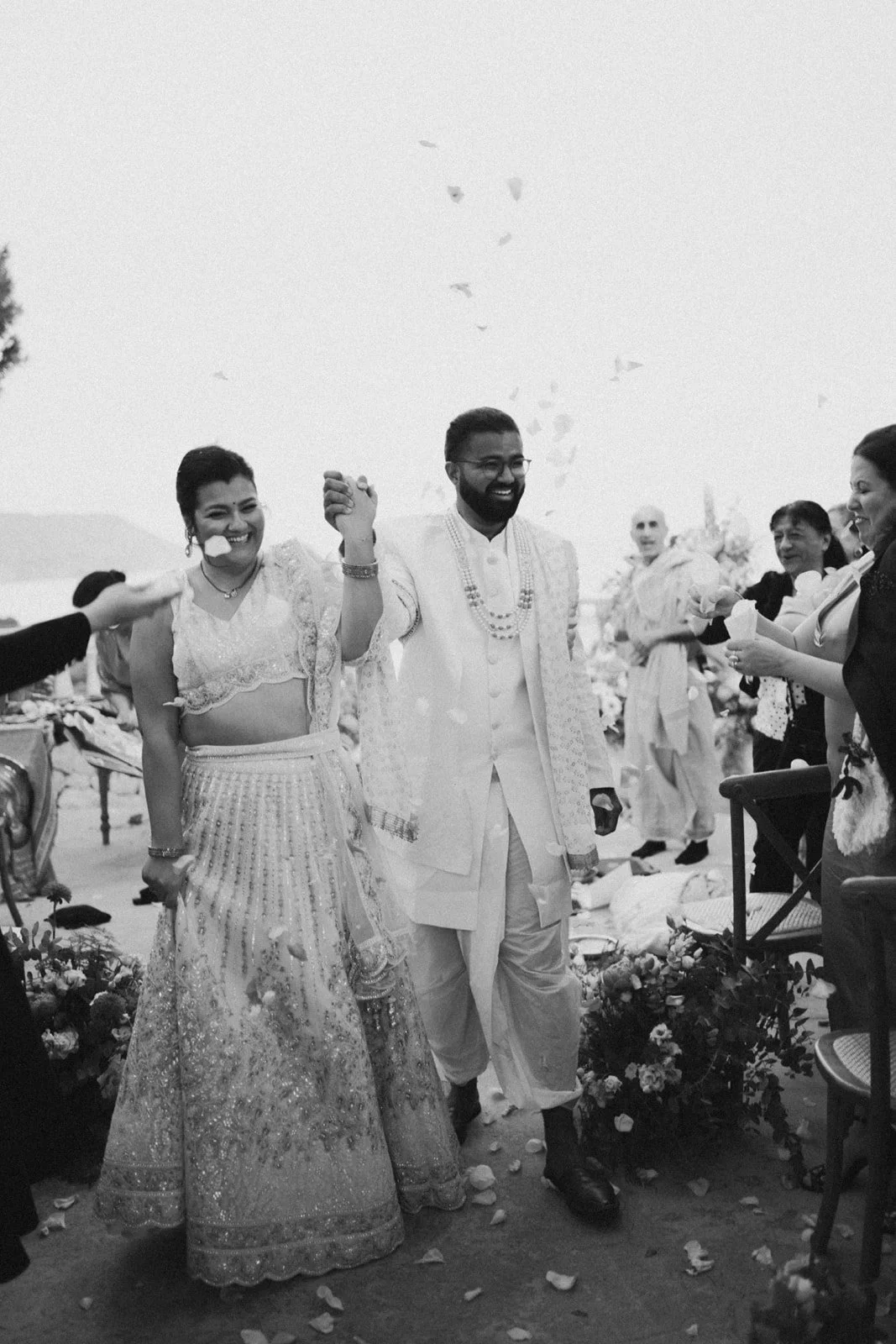 A joyful wedding celebration with a bride and groom in traditional attire, surrounded by friends and family, as flower petals are thrown in the air.