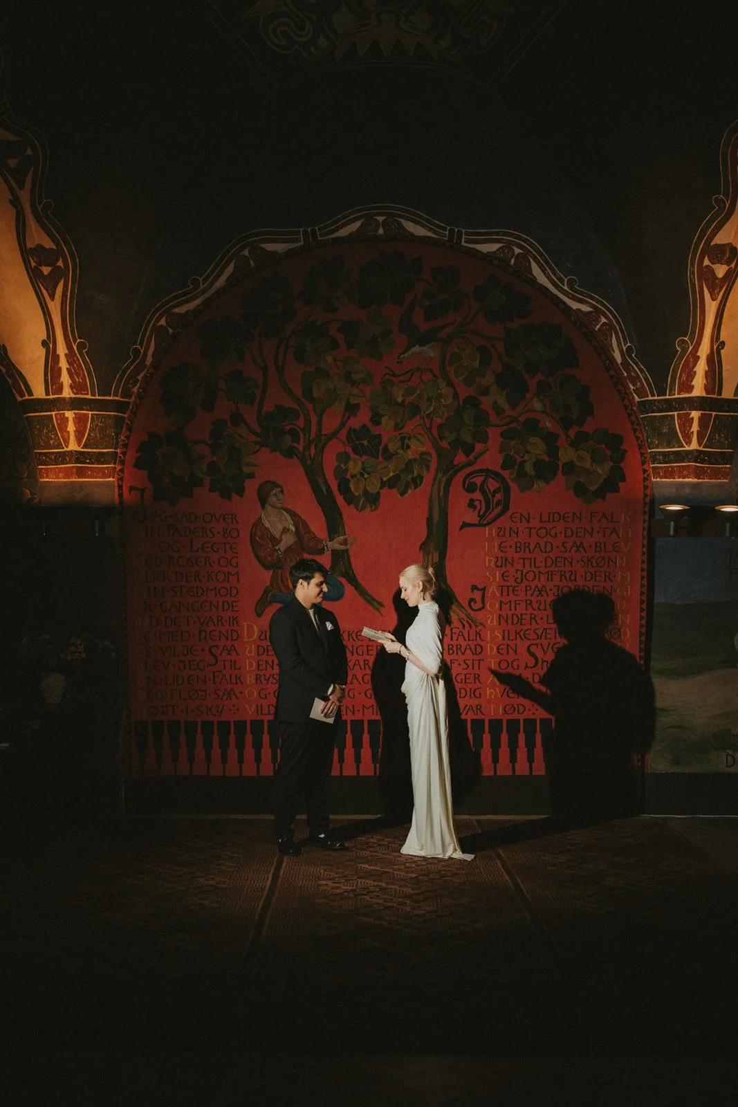 A couple standing in front of a red and gold painted backdrop with a tree and text. The man is in a black suit and the woman is in a white dress. A person in silhouette is taking a photo of them.