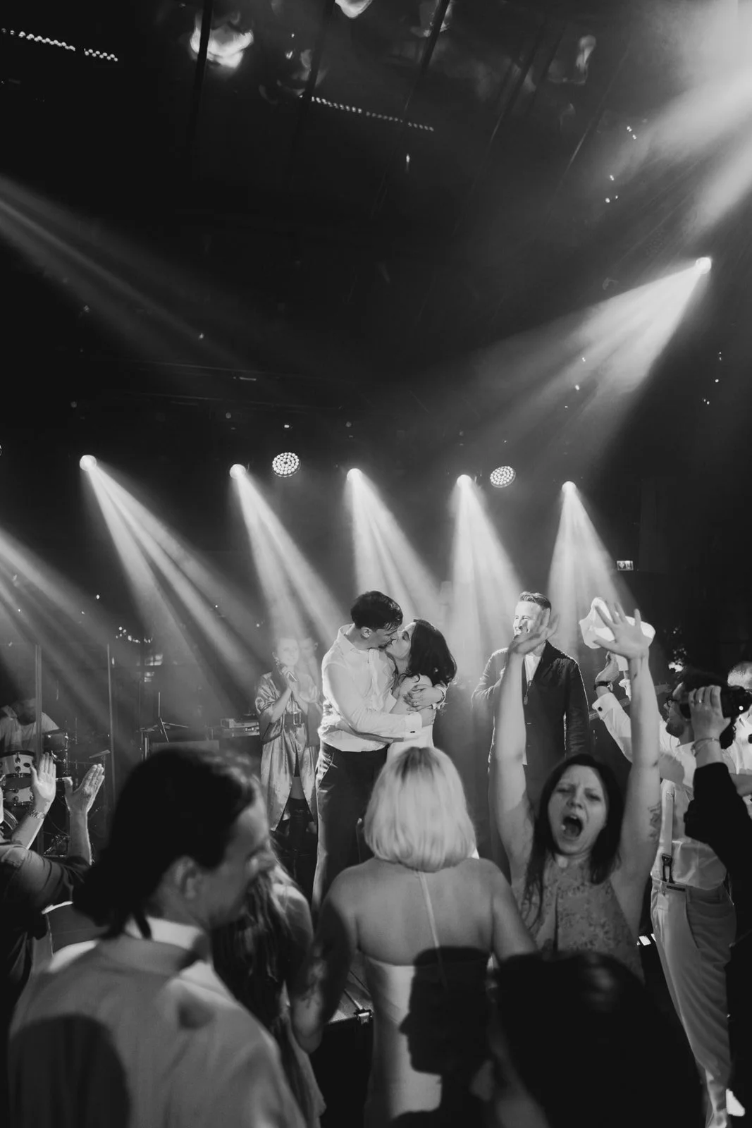 People dancing and celebrating at a party with stage lights in the background, some hugging and taking photos, in black and white.