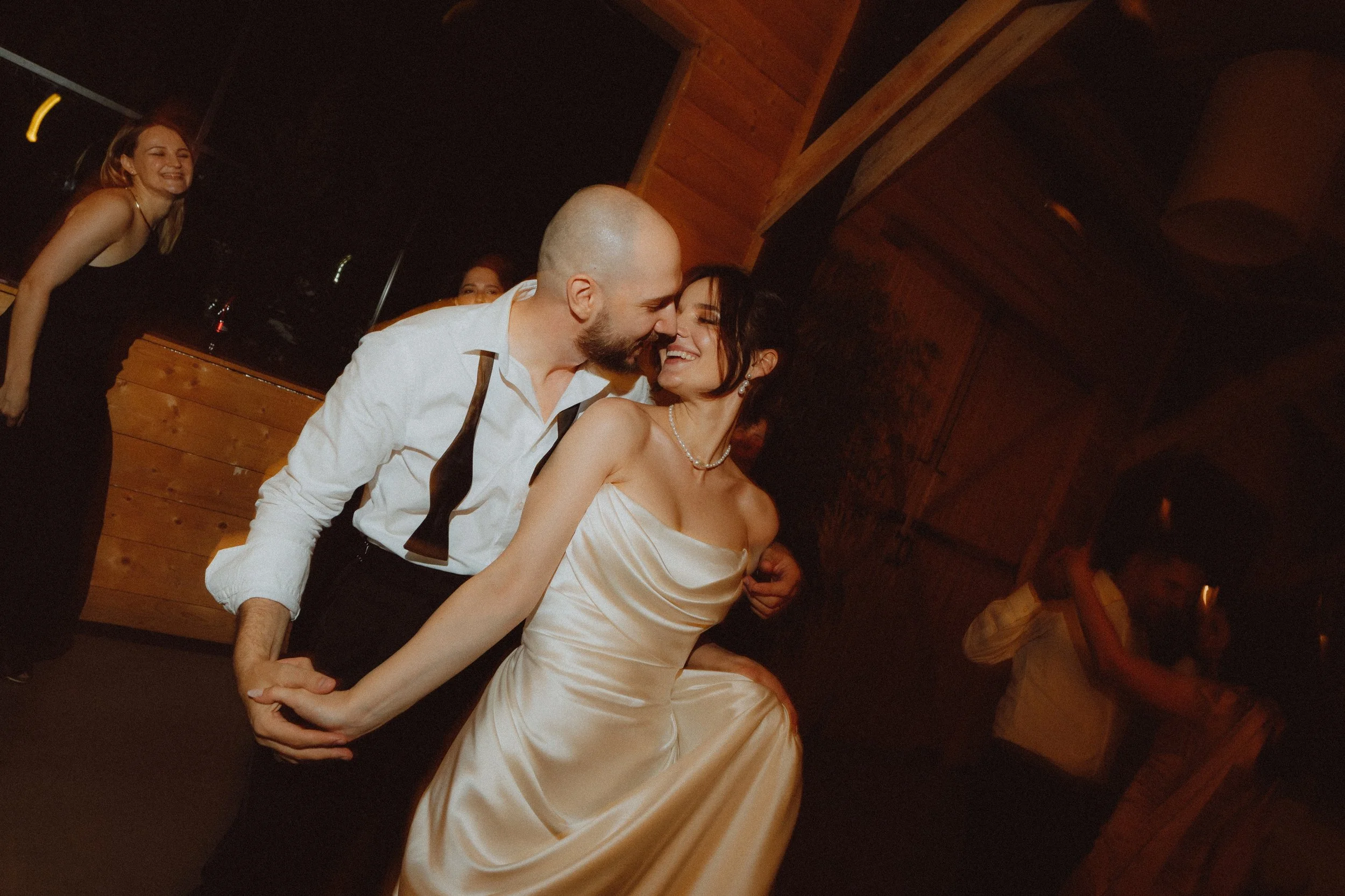 A couple dancing closely at an event, with women smiling and enjoying in the background, in a warmly lit wooden interior.
