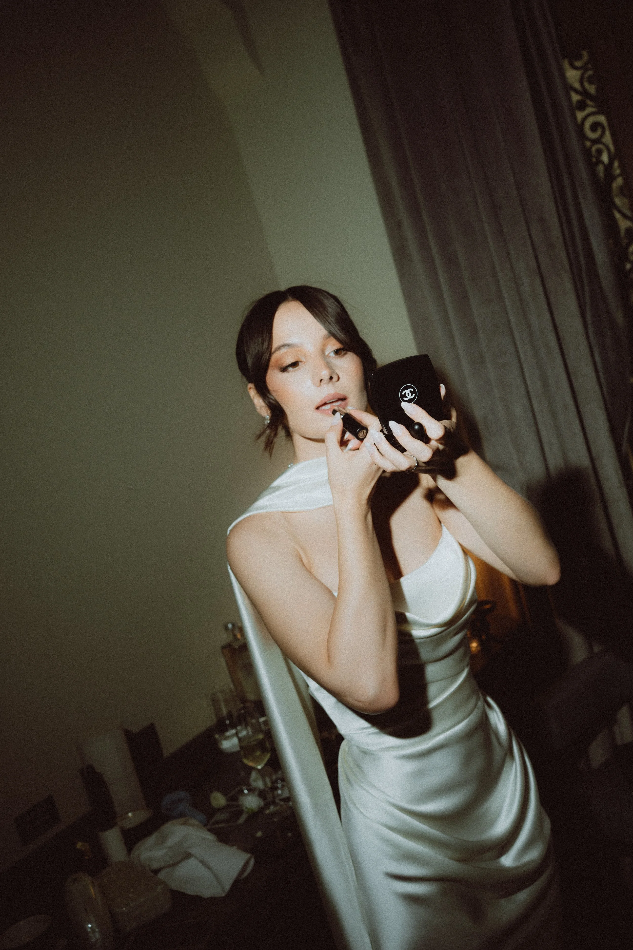 A woman with short dark hair wearing a white satin dress is looking into a small black mirror with a Chanel logo, applying lipstick.
