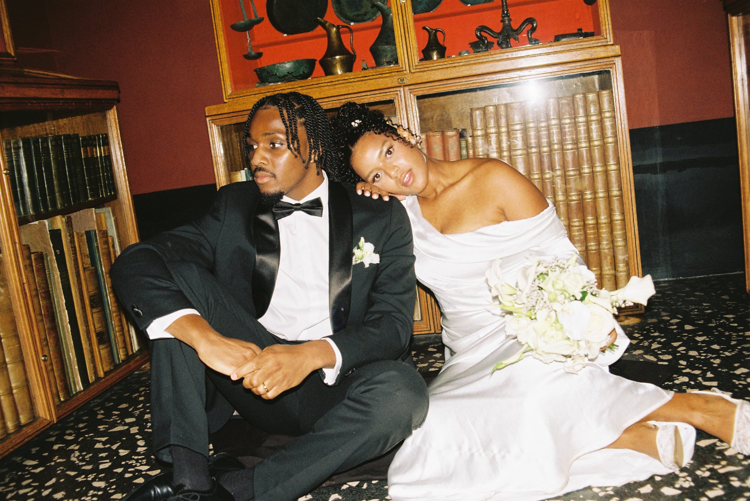 A man in a tuxedo and a woman in a wedding dress sitting on the floor with the woman leaning her head on the man's shoulder, holding a bouquet of white flowers, in a room with wooden bookshelves and decorative artifacts.
