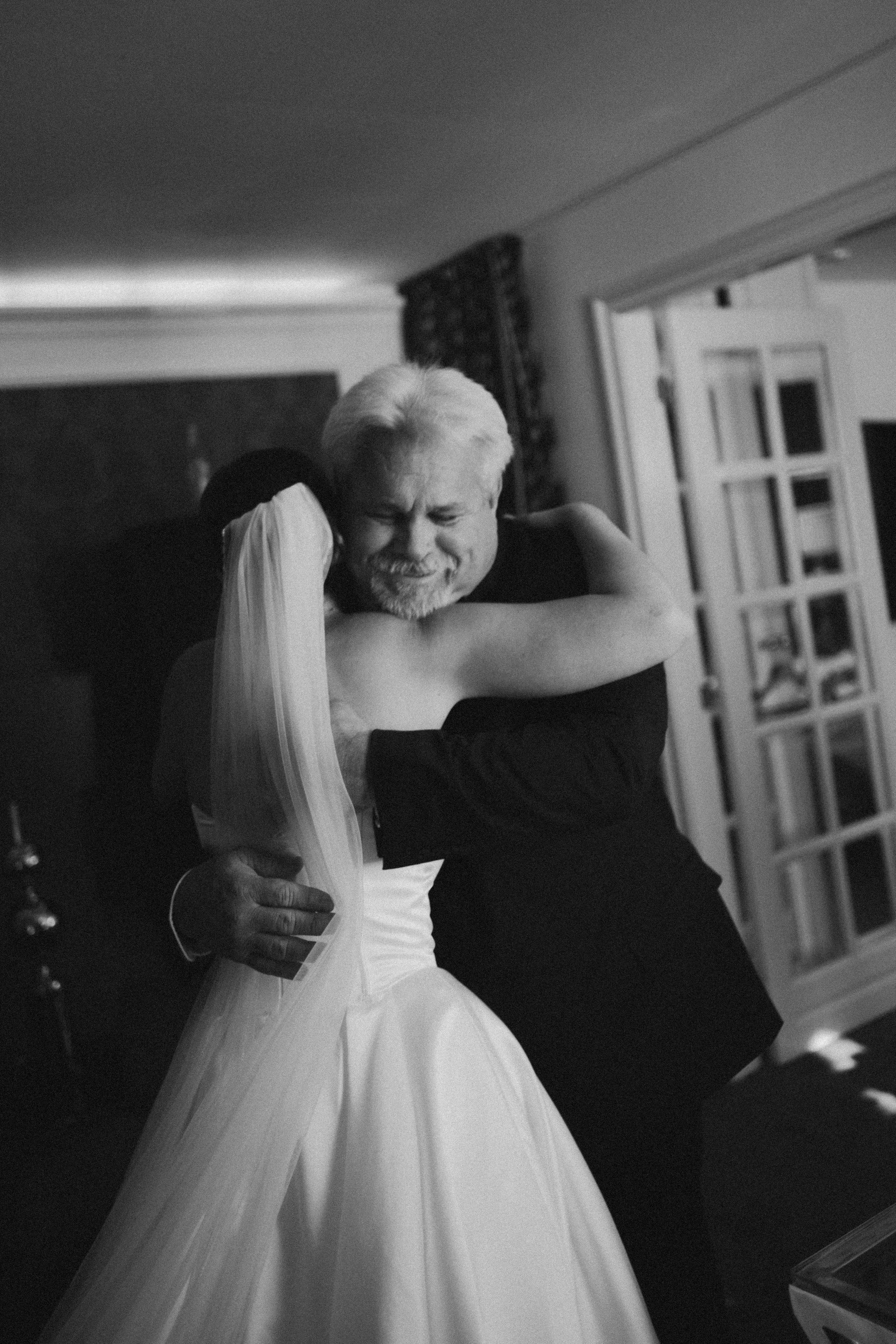 A man and a woman embrace at a wedding, with the man wearing a tuxedo and the woman in a wedding dress with a veil, smiling with eyes closed.