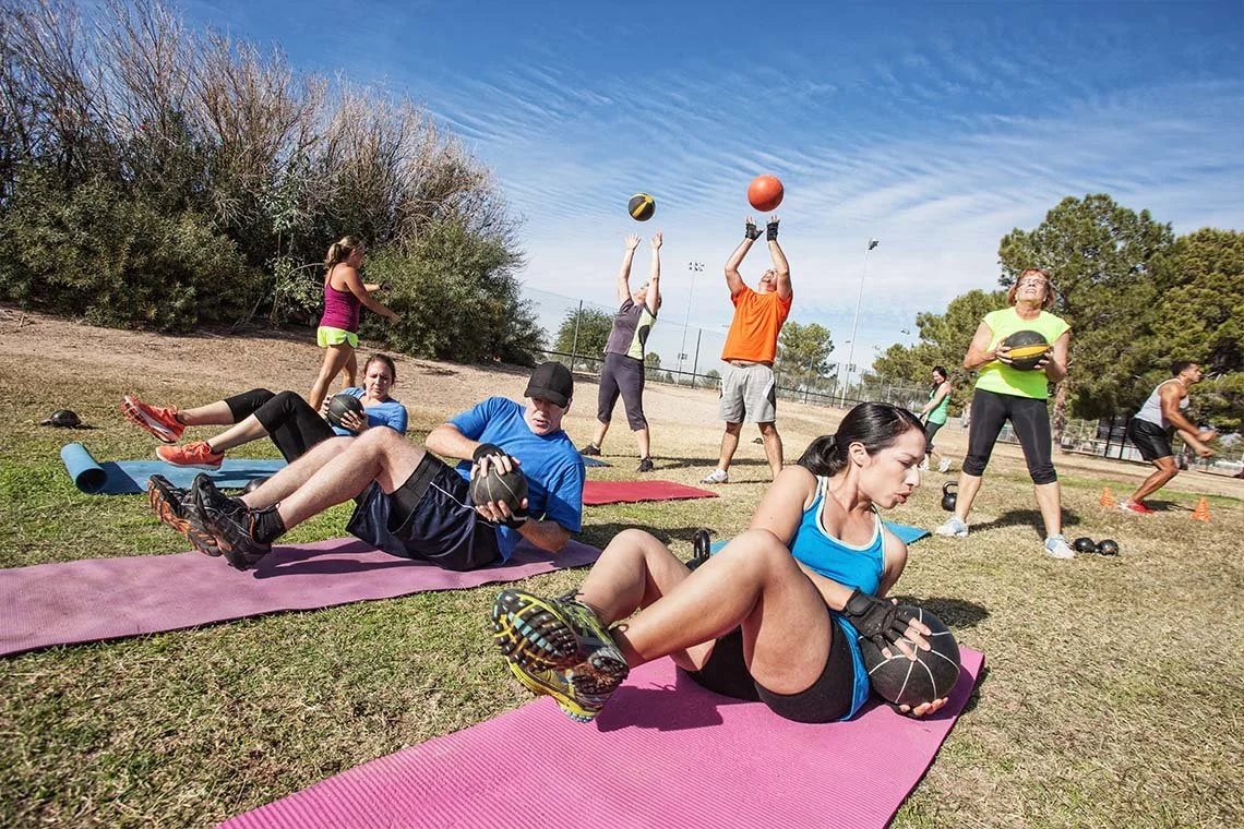 Group of people exercising outdoors on pink yoga mats, with some lifting medicine balls and others preparing for strength exercises.