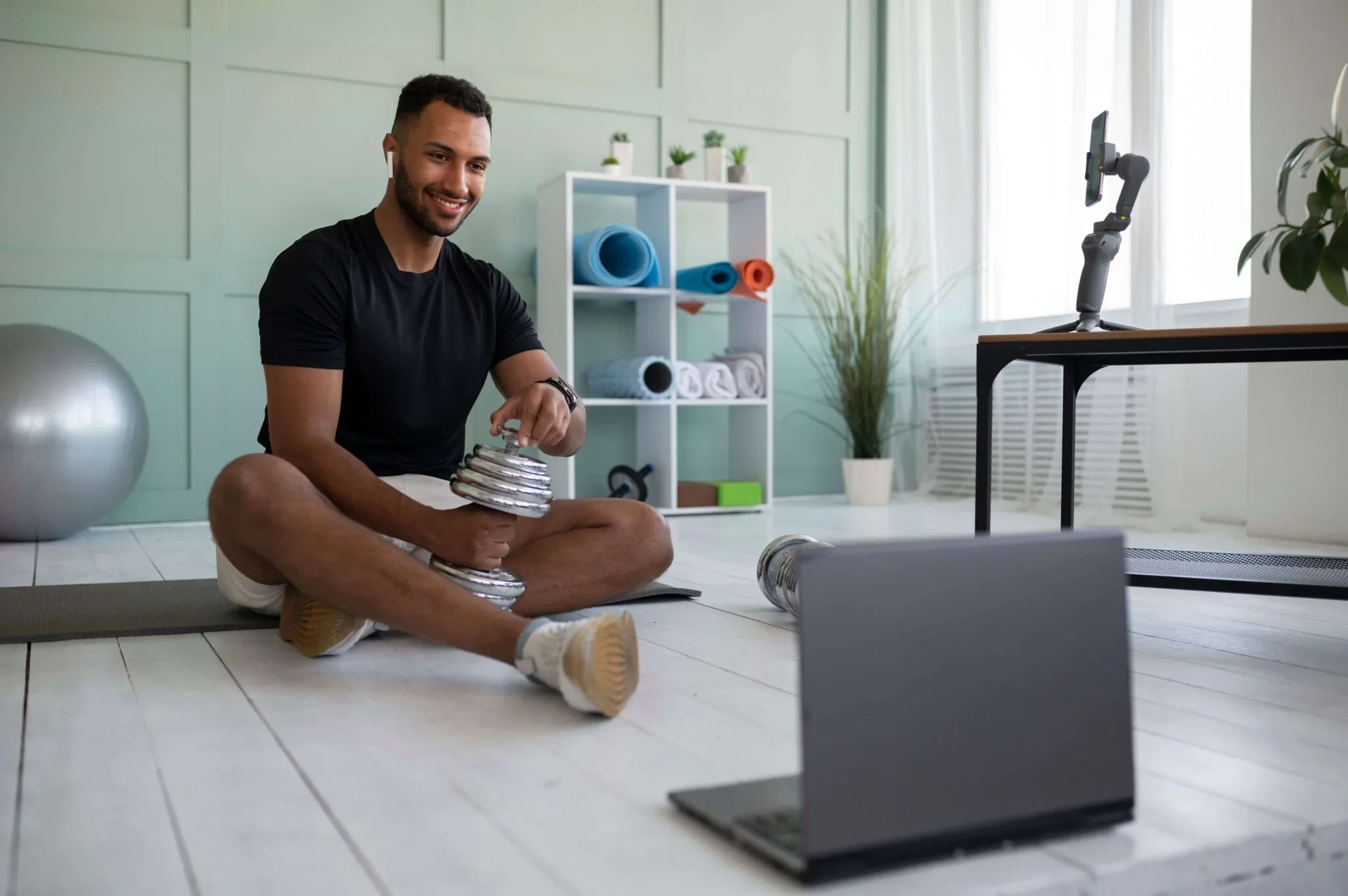 A man sitting on a yoga mat in a bright room, smiling and holding a silver dumbbell. There are workout equipment and a tablet on a table nearby, and shelves with rolled yoga mats and potted plants in the background.