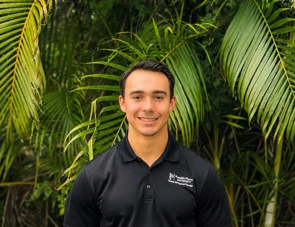 Young man with dark hair and a light complexion, smiling, wearing a black polo shirt with white logo, standing outdoors with green palm leaves in the background.