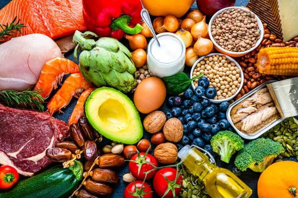A variety of fresh fruits, vegetables, nuts, grains, and dairy products arranged on a surface, including avocado, blueberries, tomatoes, broccoli, corn, eggs, milk, and various other produce.