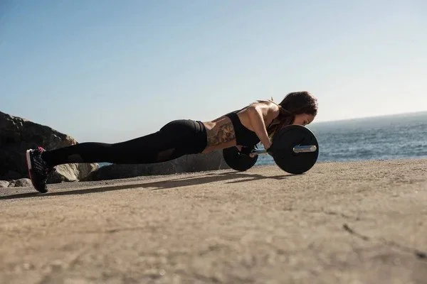 Woman doing a push-up with dumbbells outdoors on a concrete surface near the ocean.