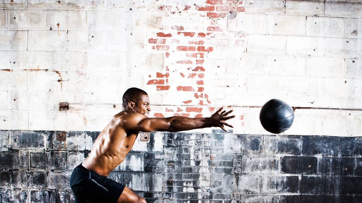 A shirtless man in black shorts throws a medicine ball against a brick wall.