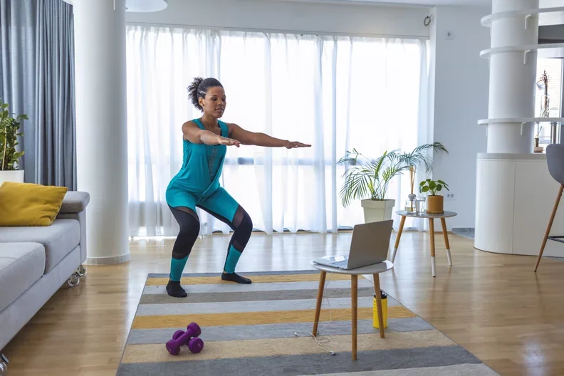 Woman in athletic wear doing squats during a virtual workout session in a bright living room, with a laptop on a small wooden table, a pair of purple dumbbells on the floor, and indoor plants in the background.