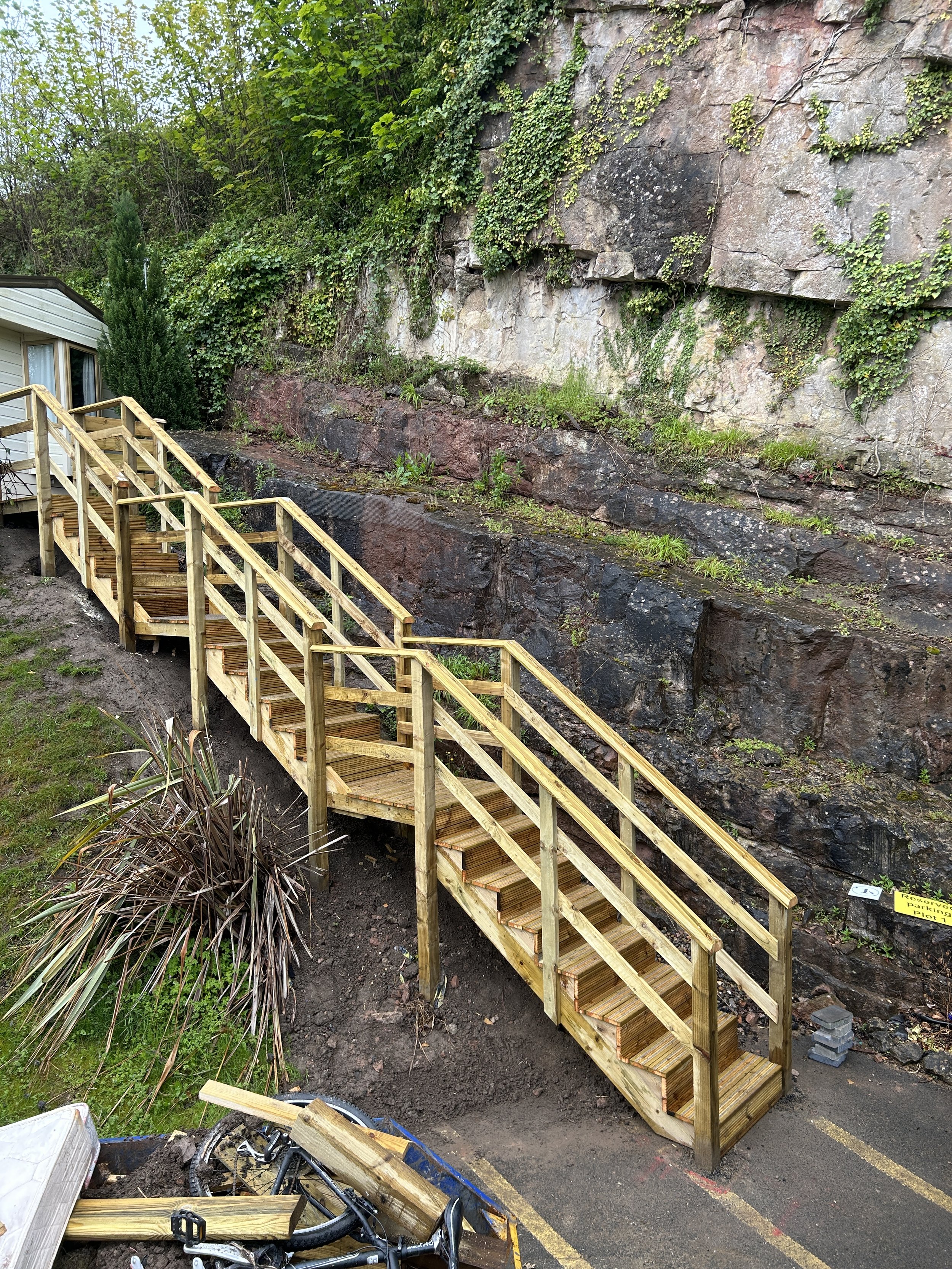 A freshly built wooden staircase with handrails runs alongside a rocky cliff face, leading up to a house with a white exterior and large windows. There are green plants and shrubs near the staircase and rocks. A small sign on the ground indicates par