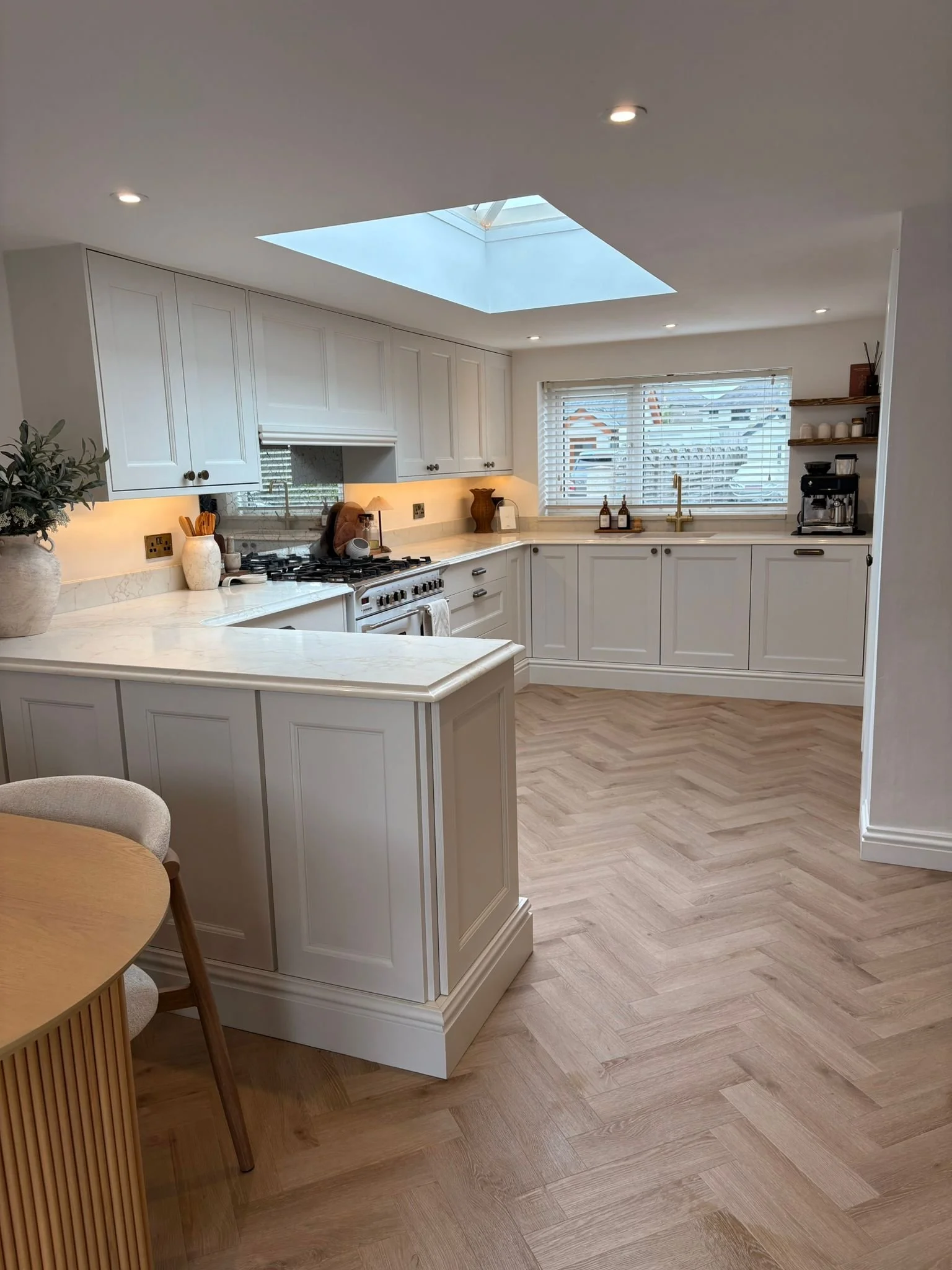 A modern kitchen with white cabinets, a skylight, a window with blinds, and a wooden floor.