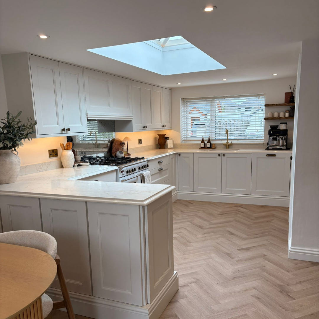 Bright kitchen with white cabinets, marble countertops, and herringbone wood flooring. Features a skylight, window with blinds, and kitchen appliances including a stove and coffee machine. Decor includes potted plant, vases, and bottles.