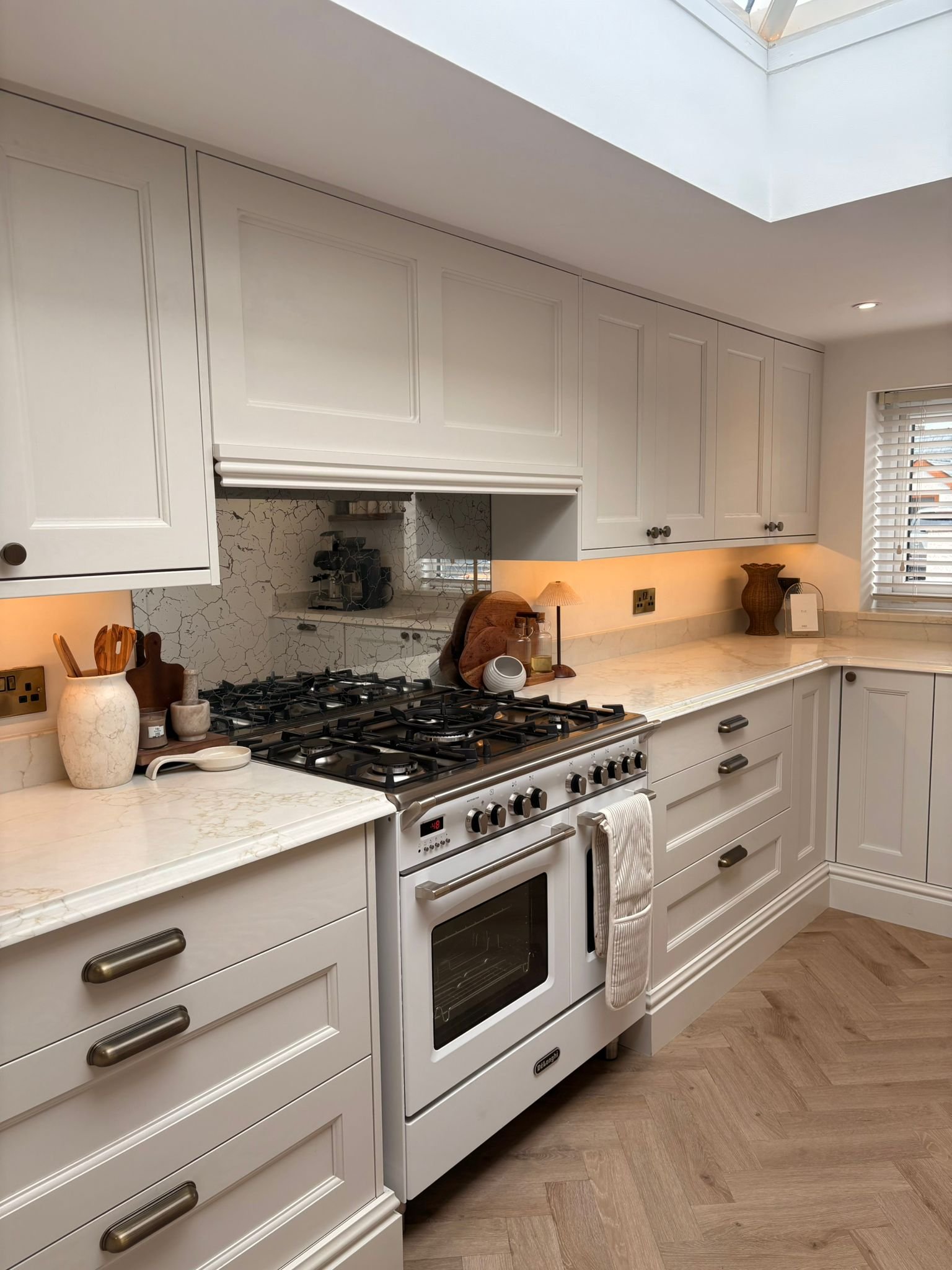A kitchen with white cabinets, marble countertops, a gas stove, and a window with blinds. There are decorative items on the counter, including a vase with wooden utensils, a small bowl, and a woven basket. The flooring is wooden in a herringbone patt