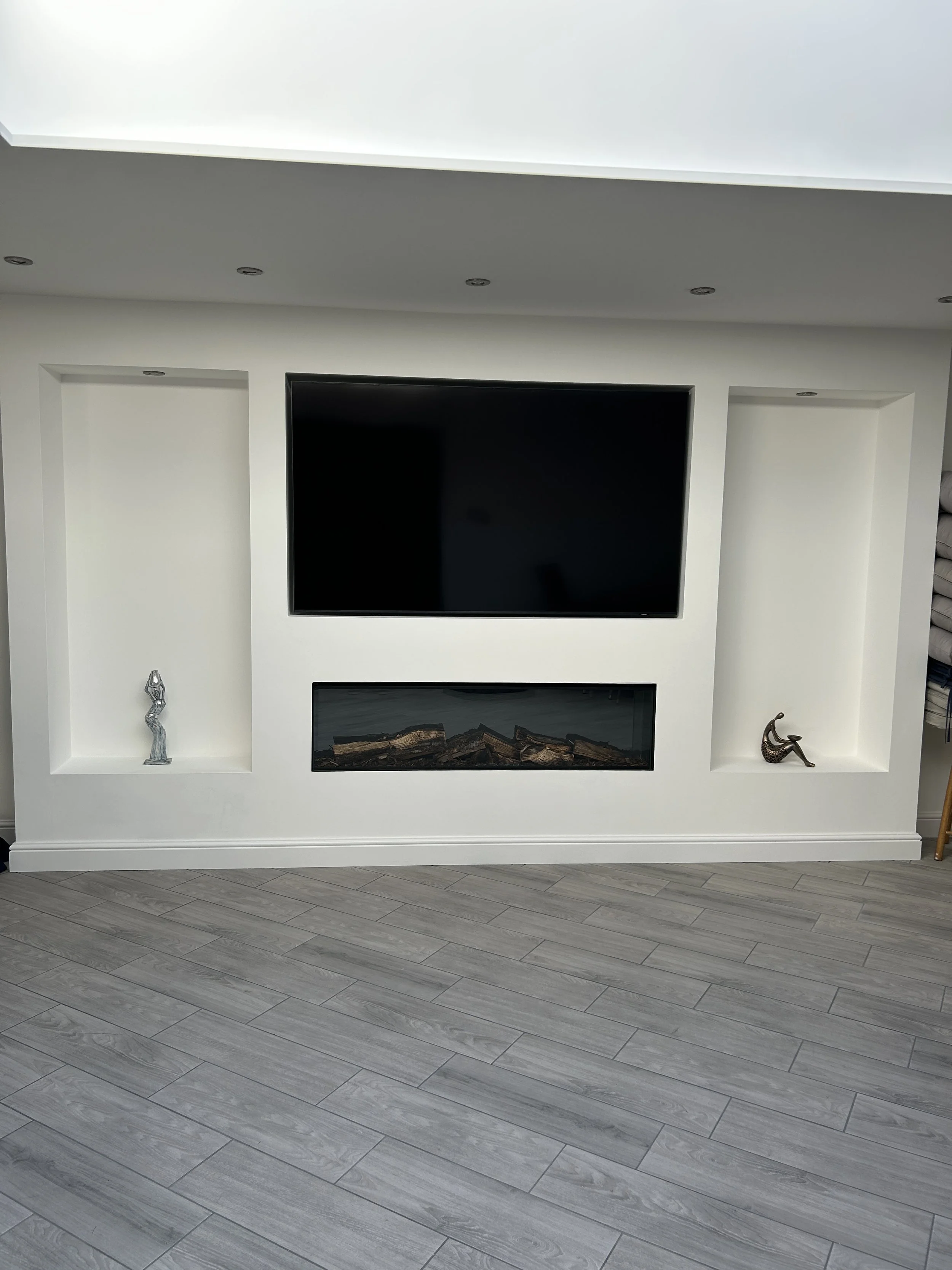 Living room wall with a mounted flat-screen TV, two recessed wall niches with sculptures, a fireplace with logs, and a gray wood floor.