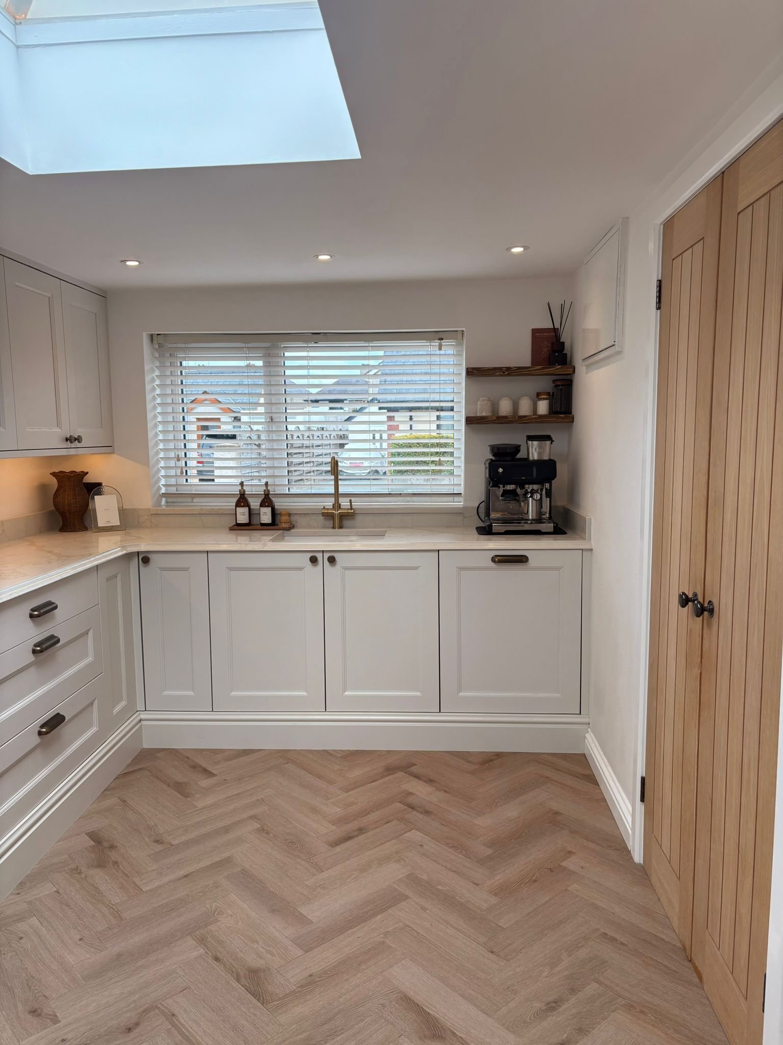 A kitchen with white cabinets, a marble countertop, a window with blinds, a coffee maker, and wooden doors on the side.