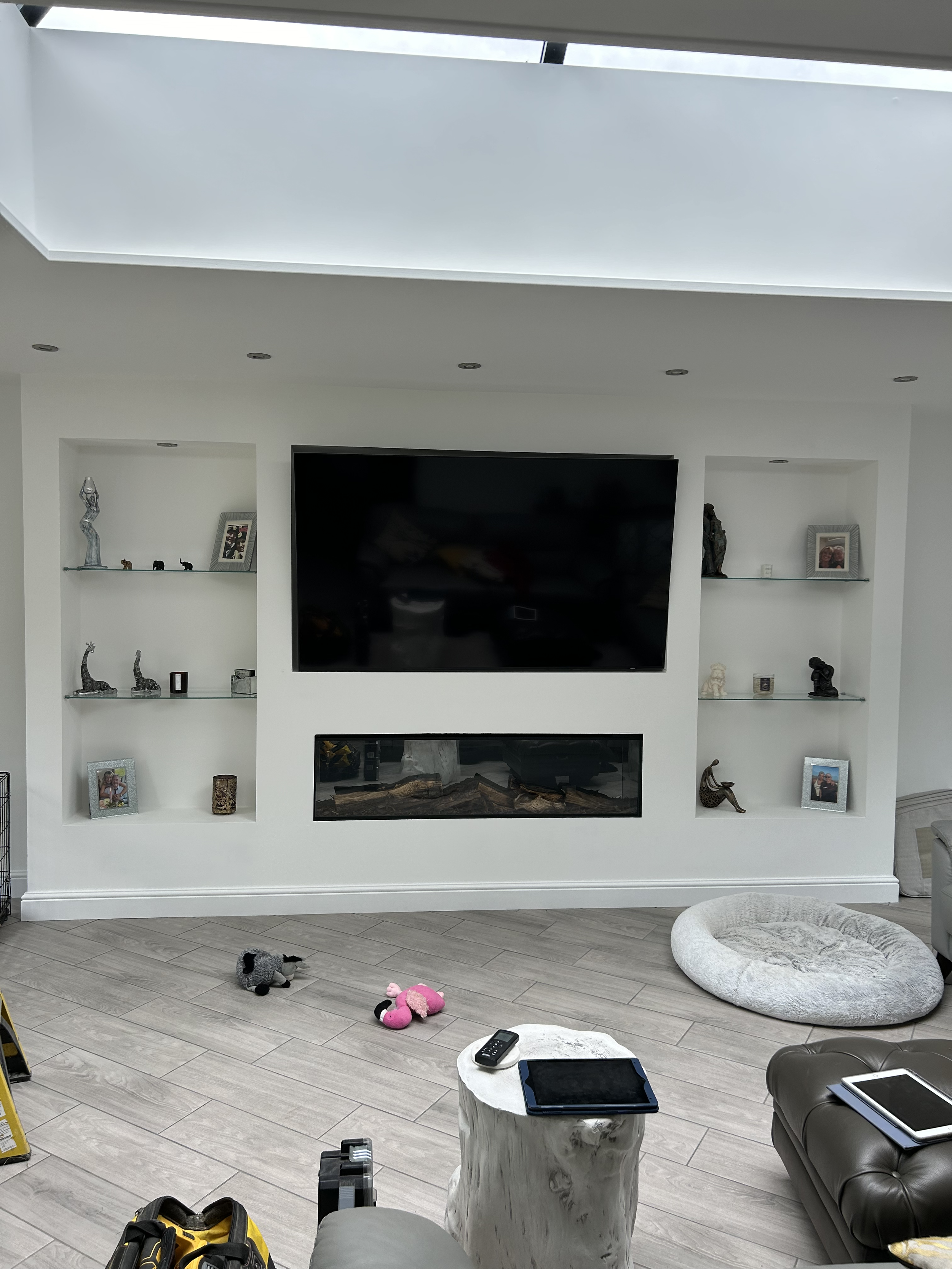 Living room with a large wall-mounted flat-screen TV, open shelves with decorative items and framed photos, a fireplace, plush pet bed, toys on the floor, and a marble side table with electronic devices.