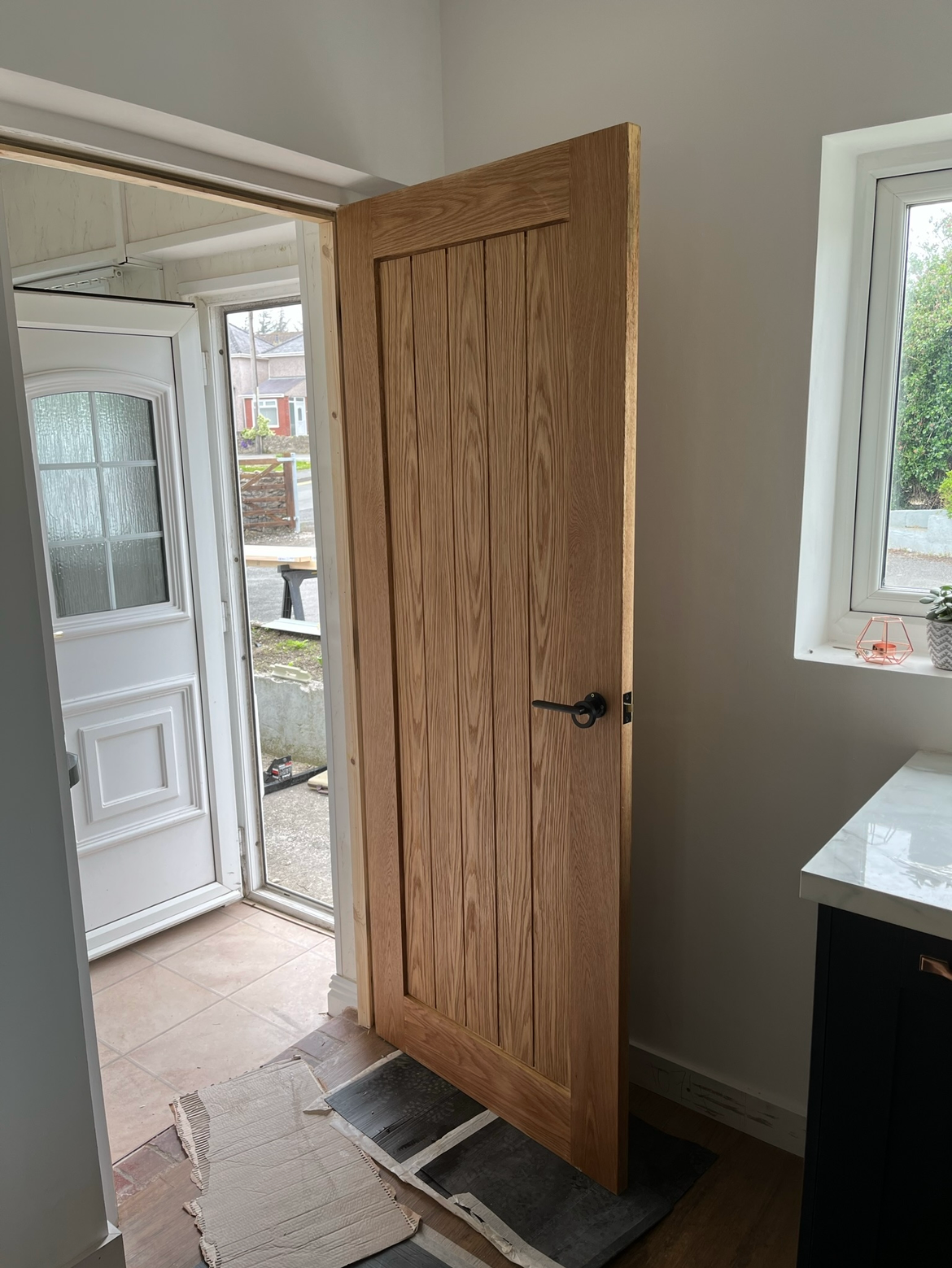 Wooden interior door partially open inside a room with a window and countertop, leading outside to a porch with a white exterior door.
