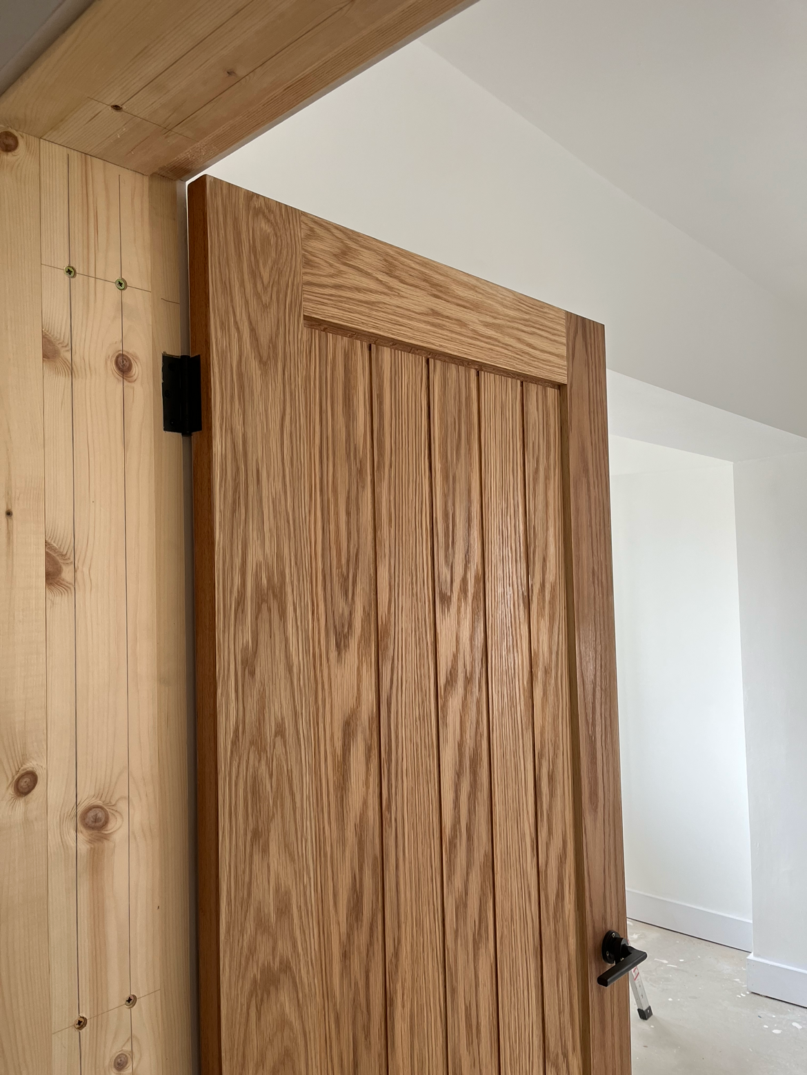 A partially opened wooden door with vertical panels, attached to a light wood door frame, in a room with white walls and a white ceiling.