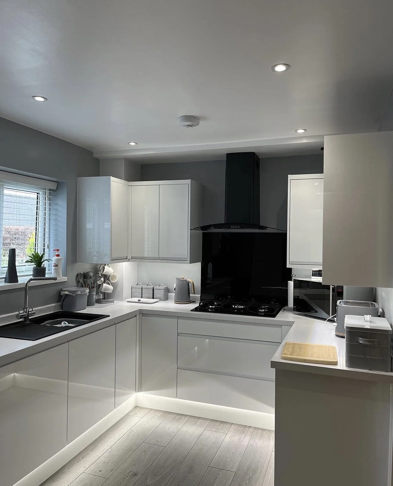 Modern white kitchen with black accents, countertop appliances, window with blinds, potted plant, and under-cabinet lighting.