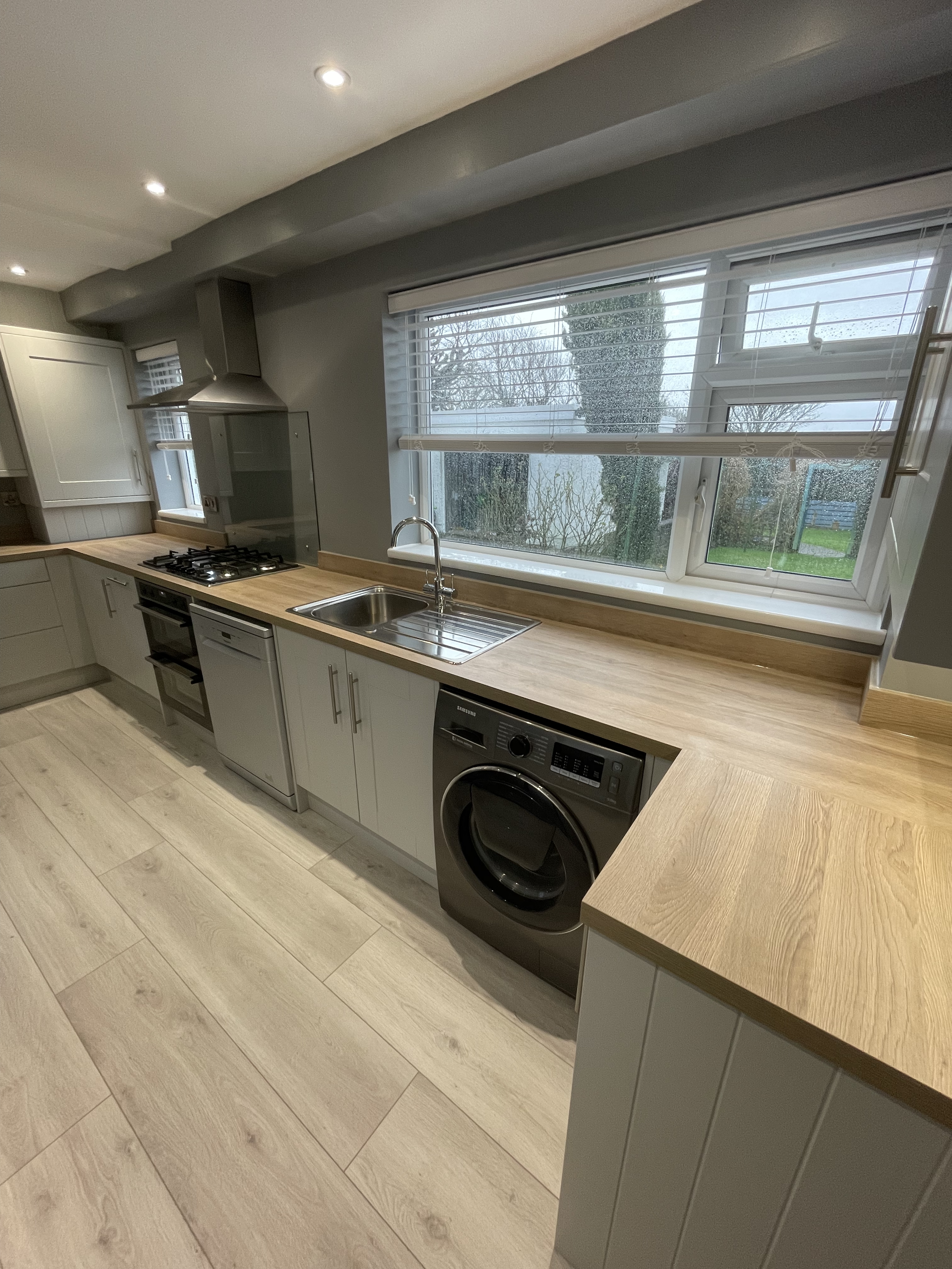 Modern kitchen featuring a wooden countertop, a stainless steel sink, a washing machine, a gas stove, and a window with blinds overlooking a rainy outdoor scene.