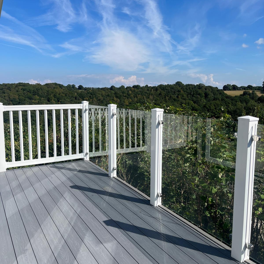 A spacious outdoor balcony with grey wooden flooring, white railings, and glass panels, overlooking a lush green landscape under a partly cloudy blue sky.