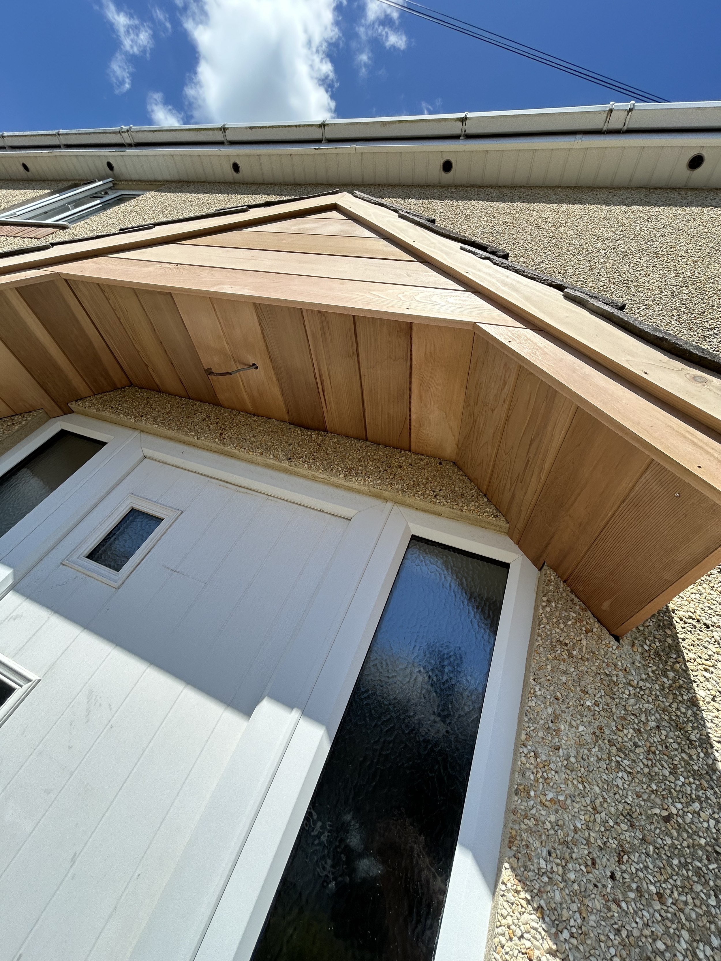 The image shows the exterior of a house with a white door, a small window, a pebble pathway, a beige textured wall, and a wooden awning with a black drainage channel beneath it. The sky is partly cloudy with a blue backdrop.