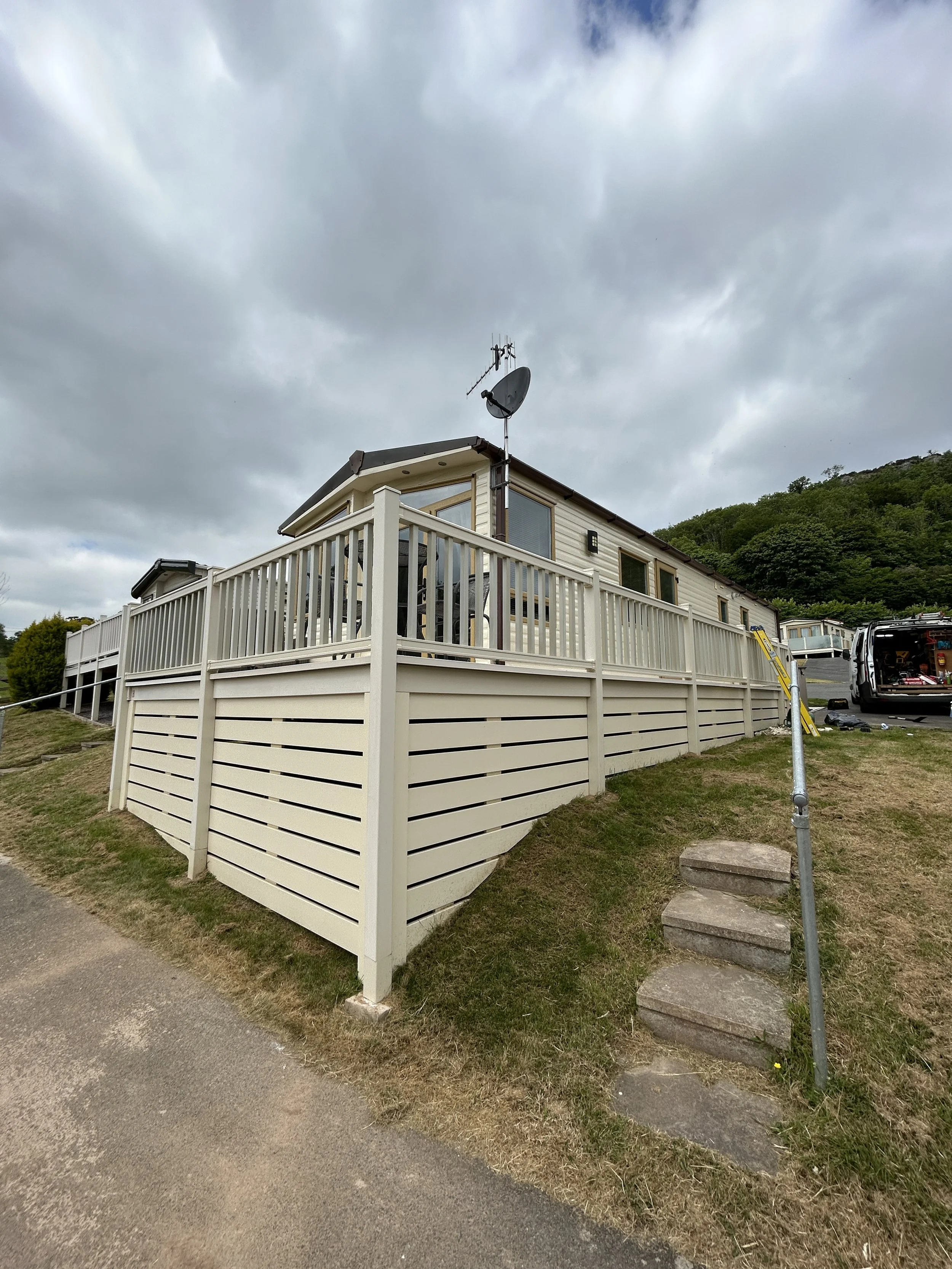A two-story mobile home with a spacious balcony, satellite and TV antennas on roof, and a white fence surrounding the yard. Overcast sky and green hillside in background.