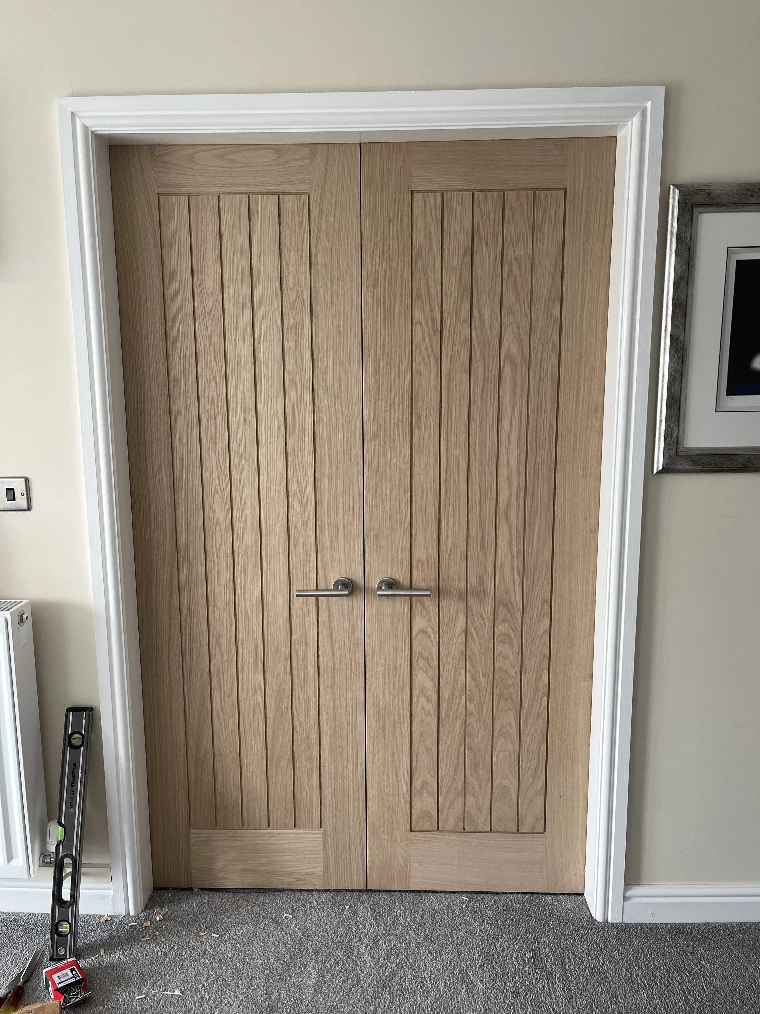 Wooden double doors with silver handles, white trim, beige walls, gray carpeted floor, level tool on the left, and a framed picture on the right.