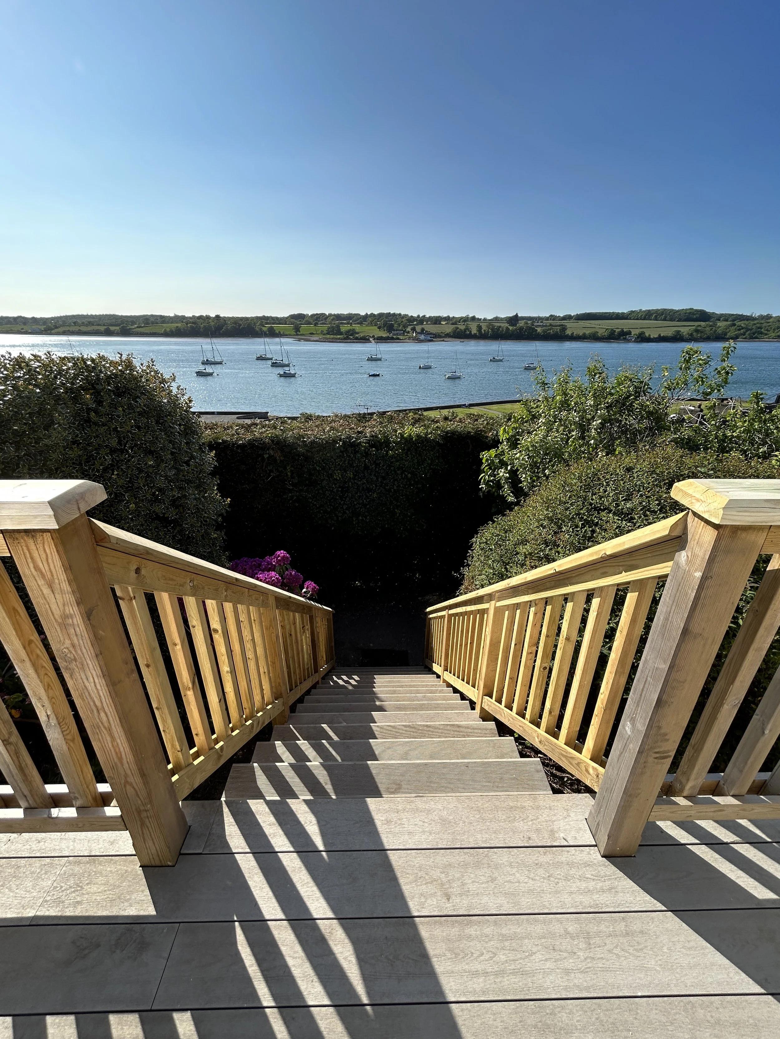 Wooden stairs leading down to a view of a body of water with sailboats, greenery, and blue sky in the background.