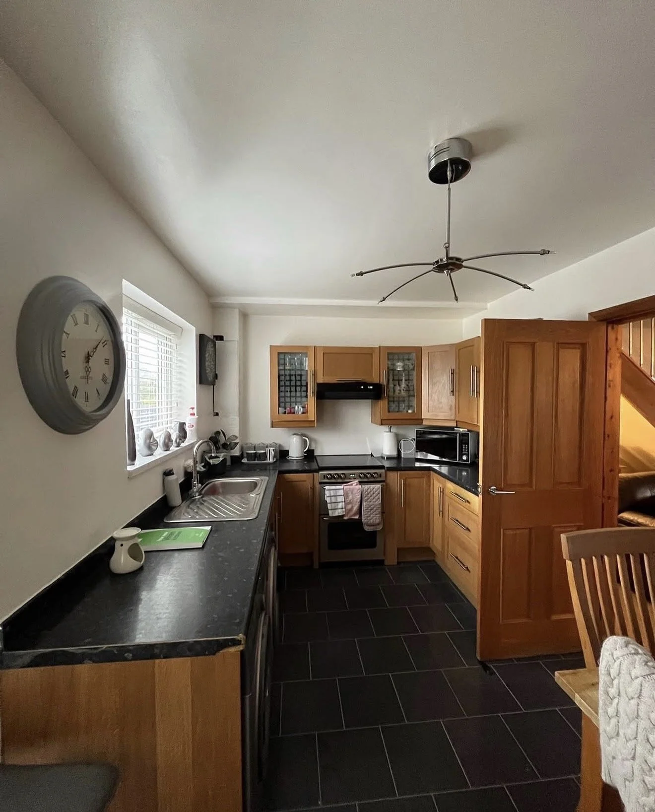 A kitchen with wooden cabinets, a black countertop, and black tiled floor. There is a clock on the wall, a sink under a window, and various appliances including a microwave and a stove.