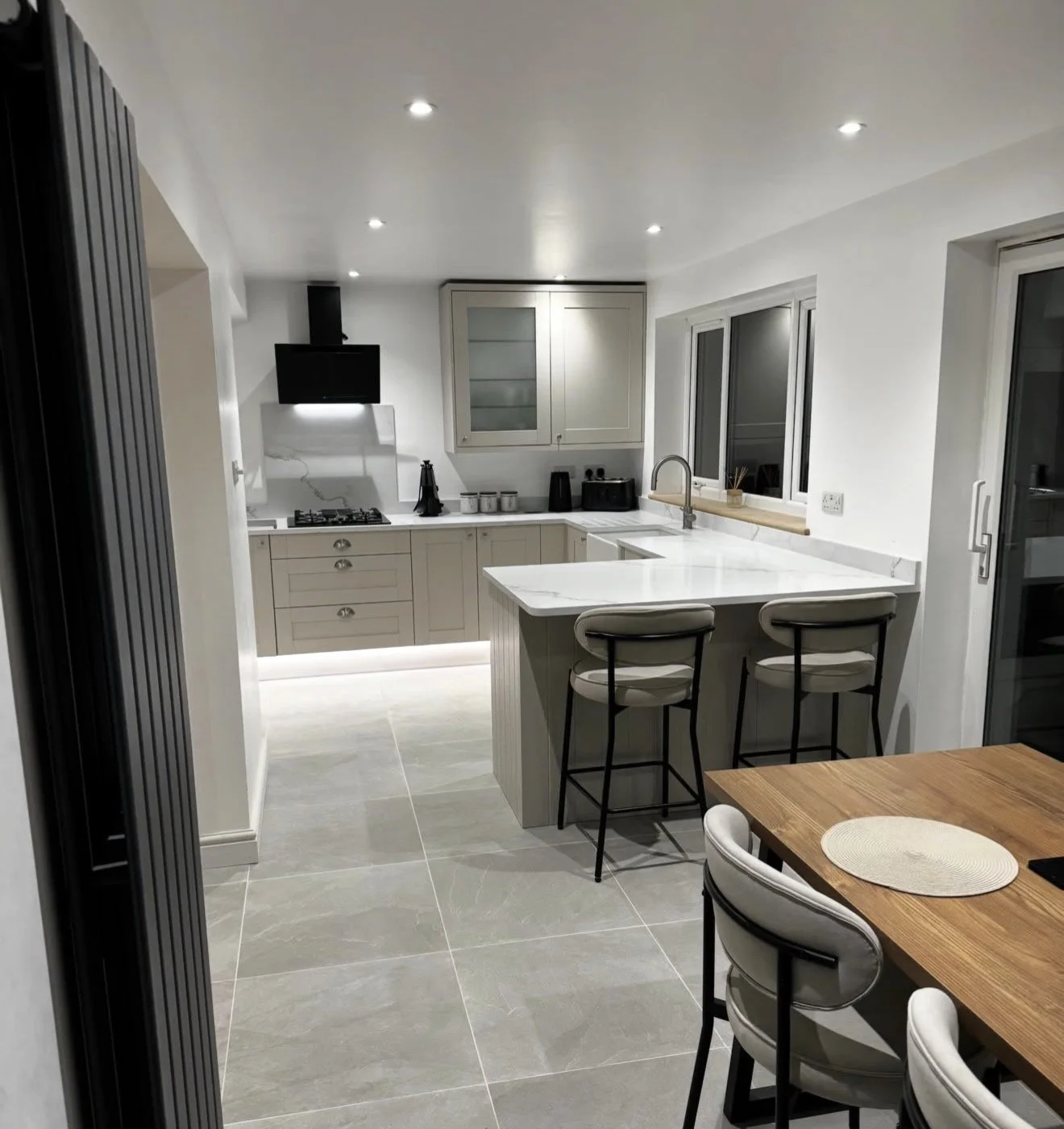 Modern kitchen with white cabinets, grey countertops, and a marble island with barstools. Includes a tv, stove, and large window with black frames.