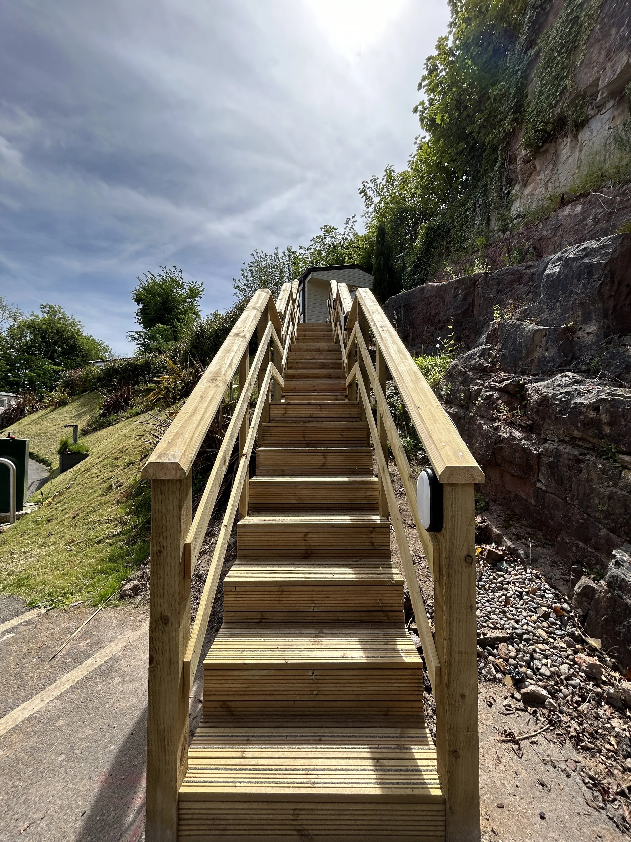 Wooden staircase leading up a hill, with a railing on both sides, alongside a rocky cliff, against a cloudy sky.