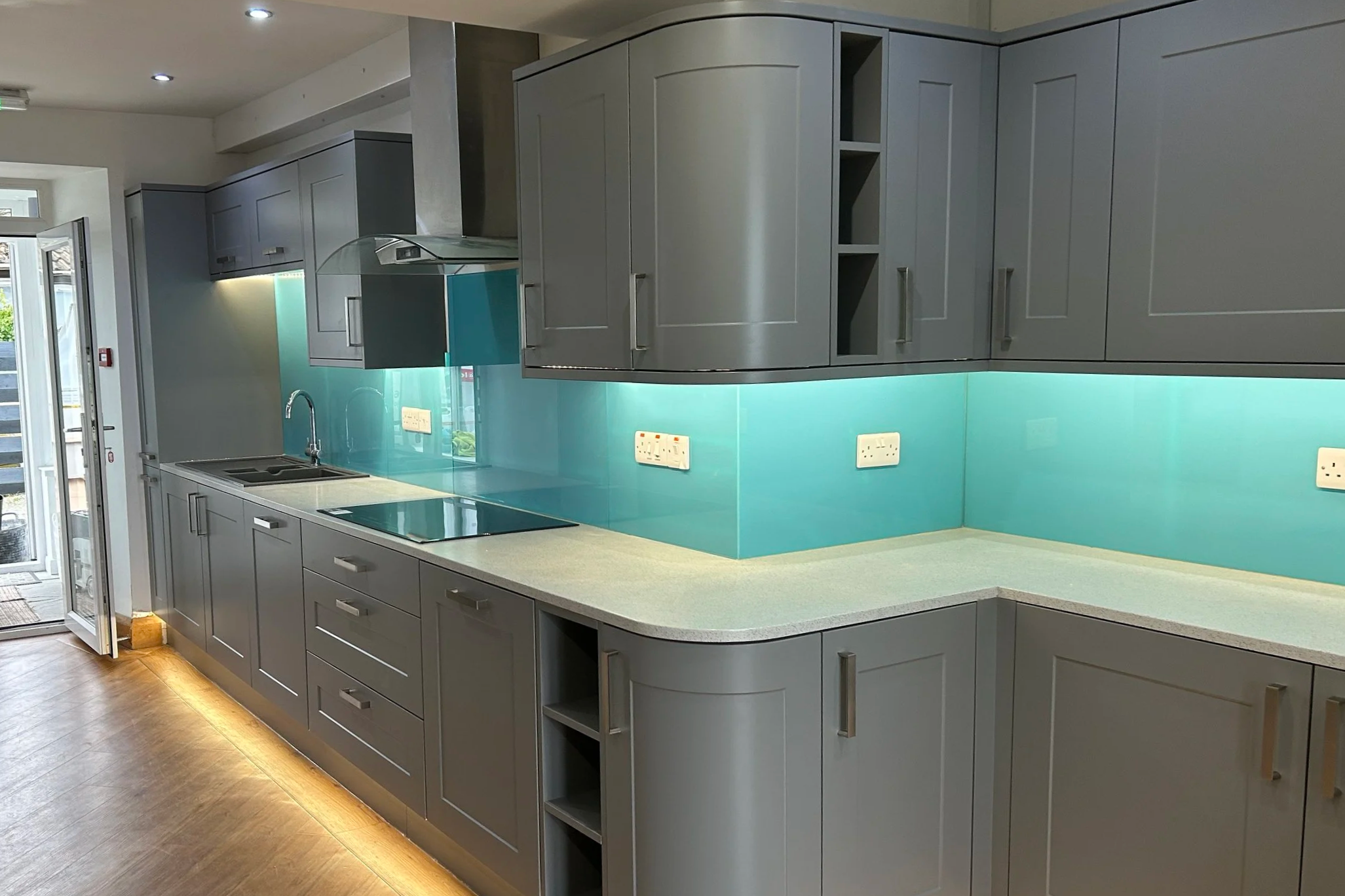 Modern kitchen with gray cabinets, a gray countertop, and a light blue glass backsplash. There is a sink with a faucet, electrical outlets on the backsplash, and a door leading outside on the left. Recessed ceiling lights illuminate the space.