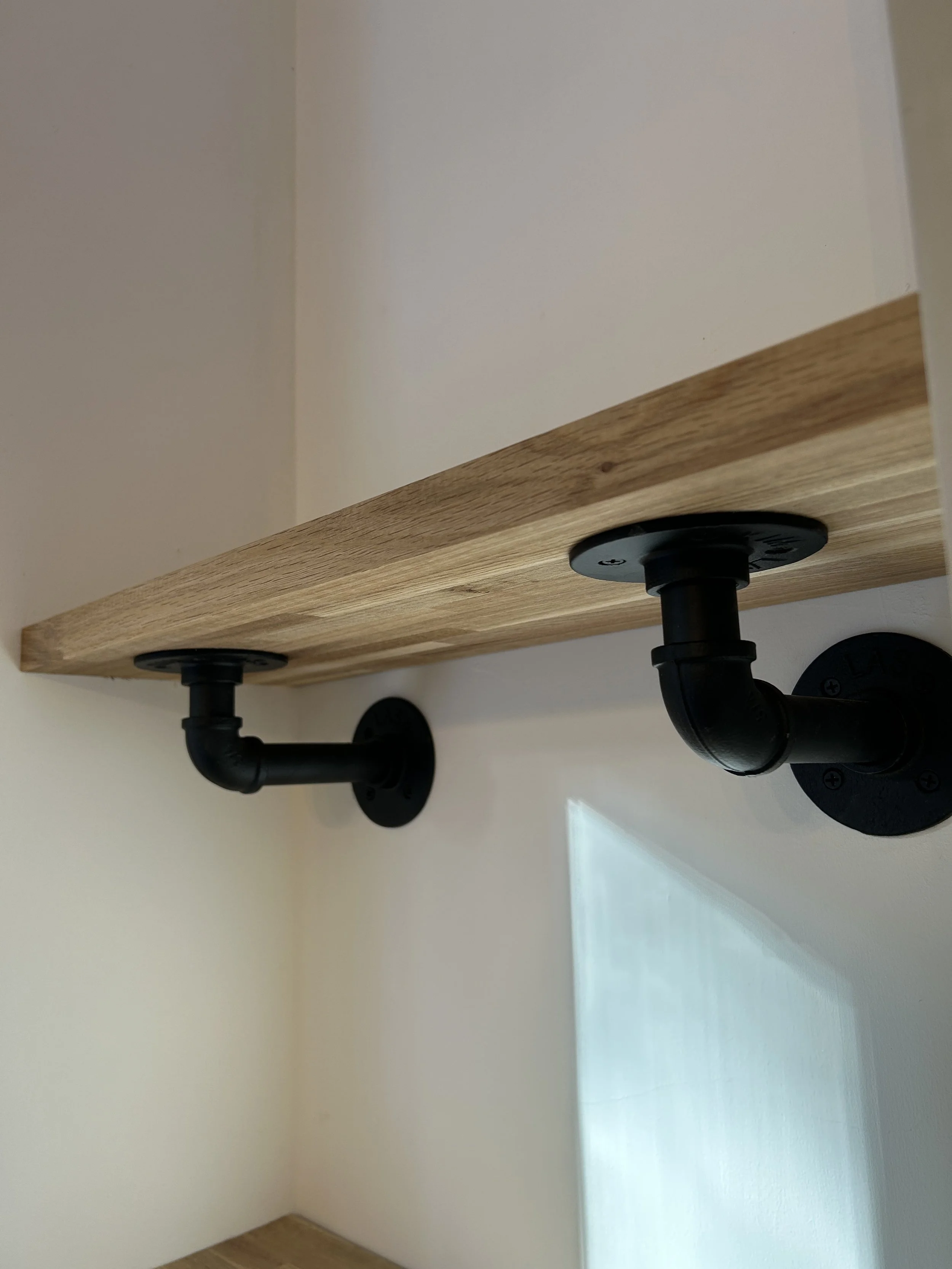Close-up of a wooden shelf attached to the wall with black metal brackets and piping underneath.