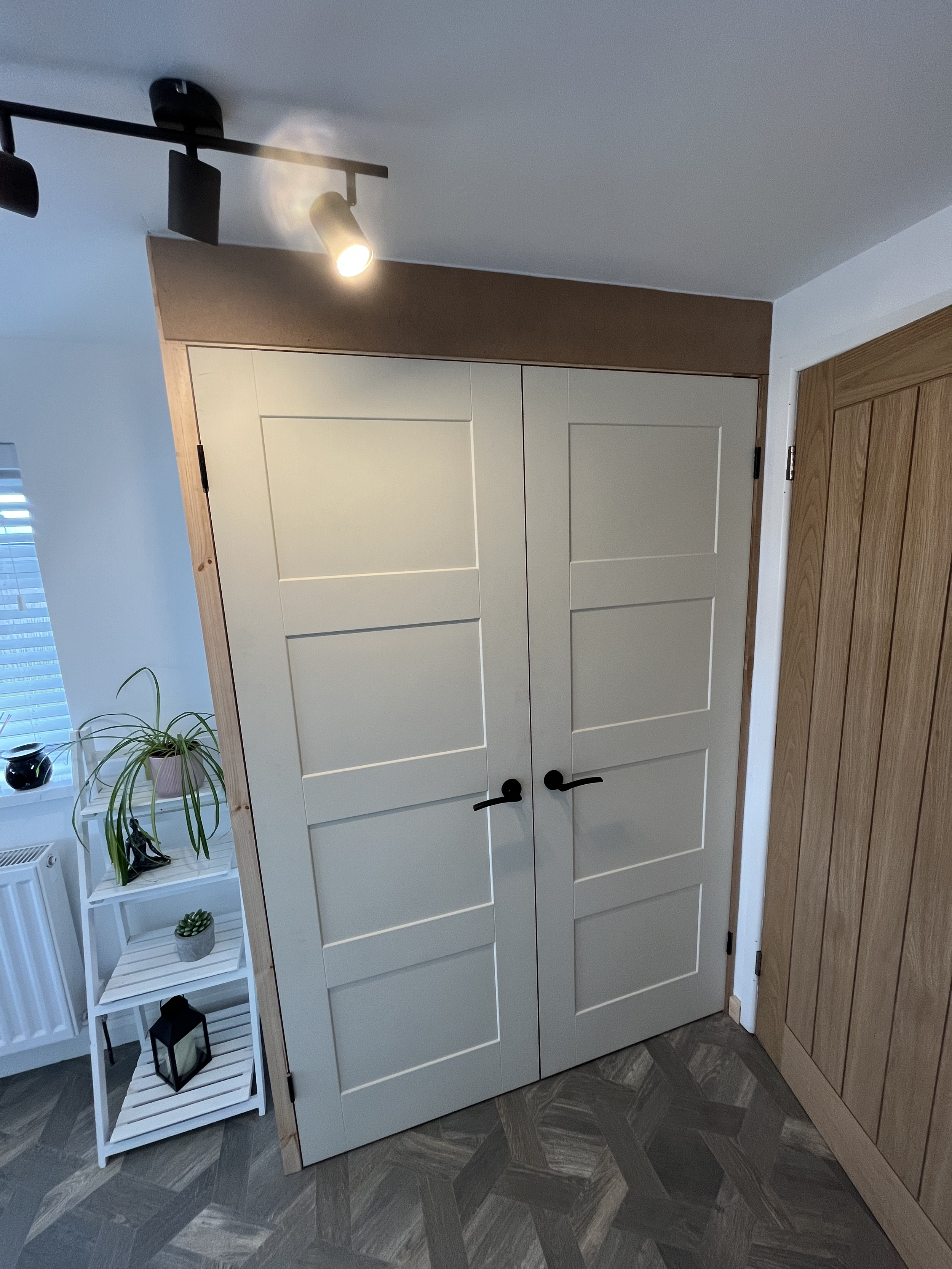 White cabinet with doors, wooden trim, and black handles in a room with gray tile flooring, next to a wooden door with panels and a white shelving unit holding plants and decor items.