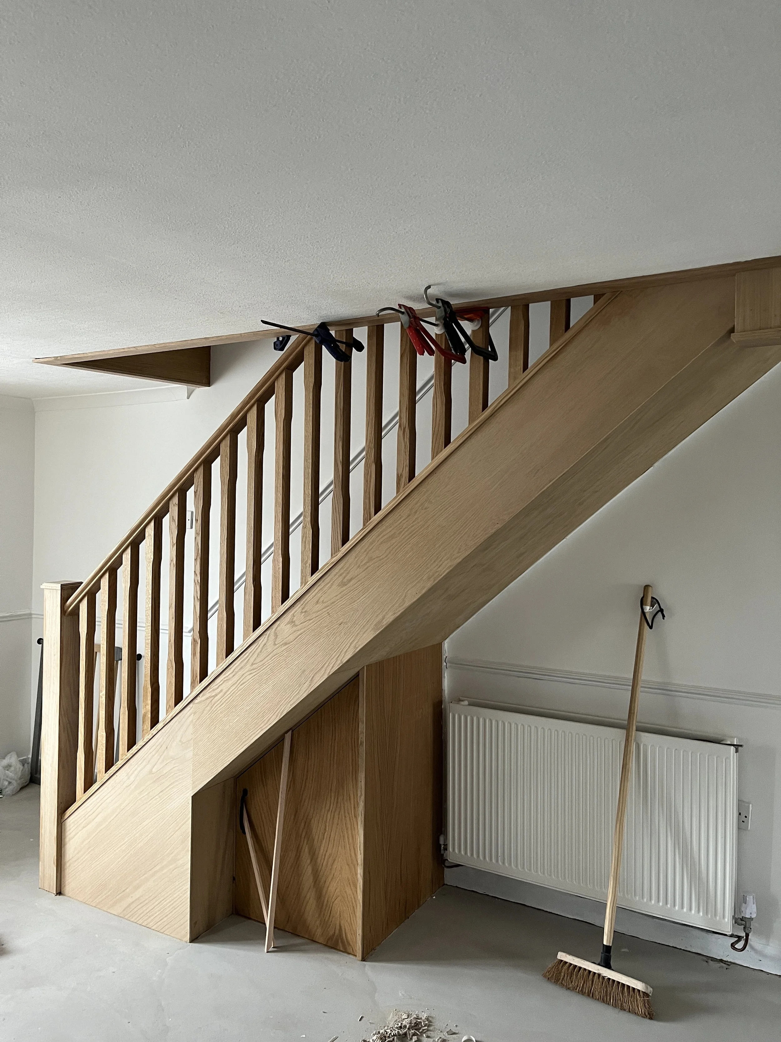 Unfinished wooden staircase with handrails, cleaning tools including a broom and trash on the floor, and a radiator against a white wall in a room under construction.
