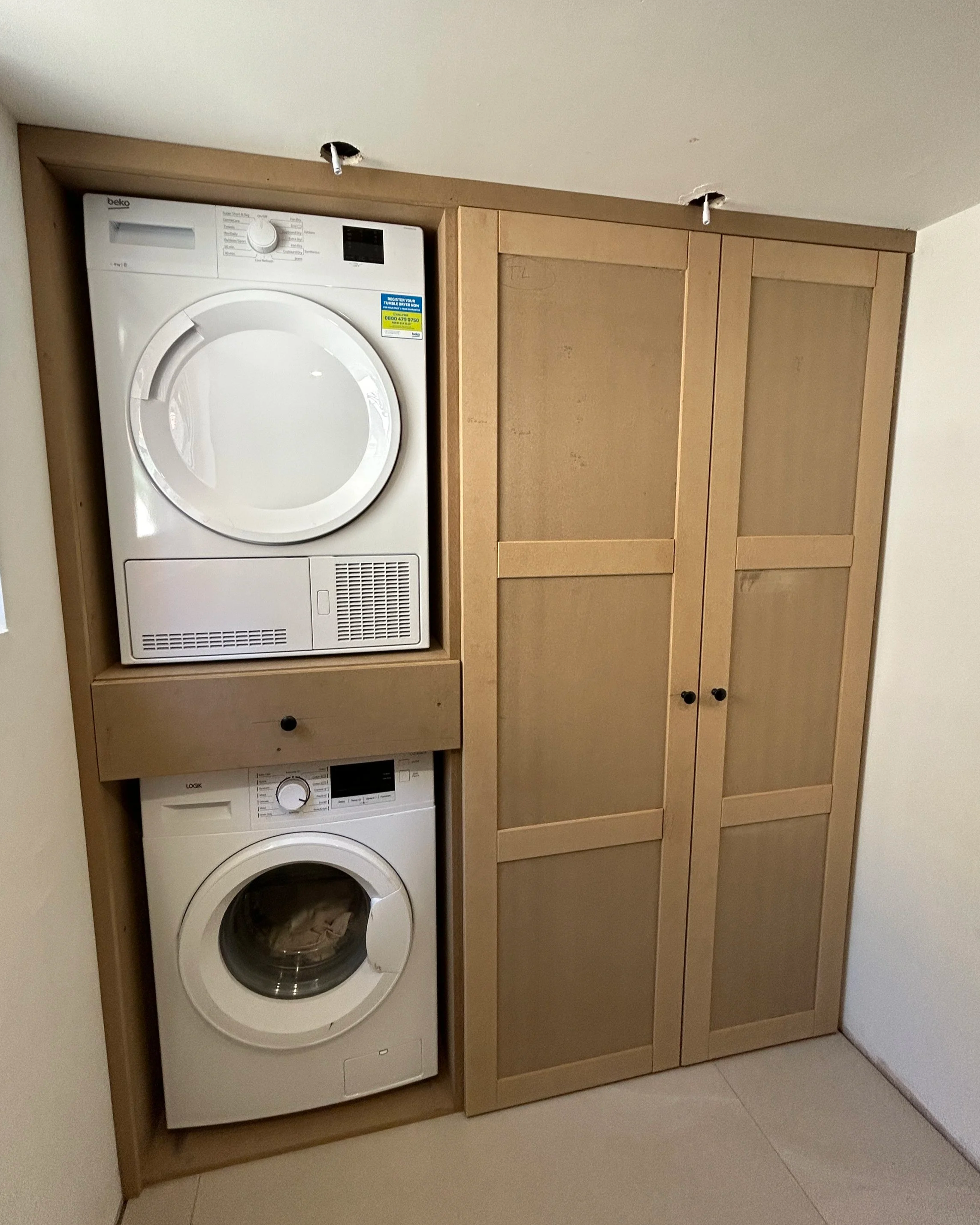 A laundry area with a white washing machine and dryer stacked, enclosed in a light wood cabinet with sliding doors, located in a corner with a light floor and plain walls.