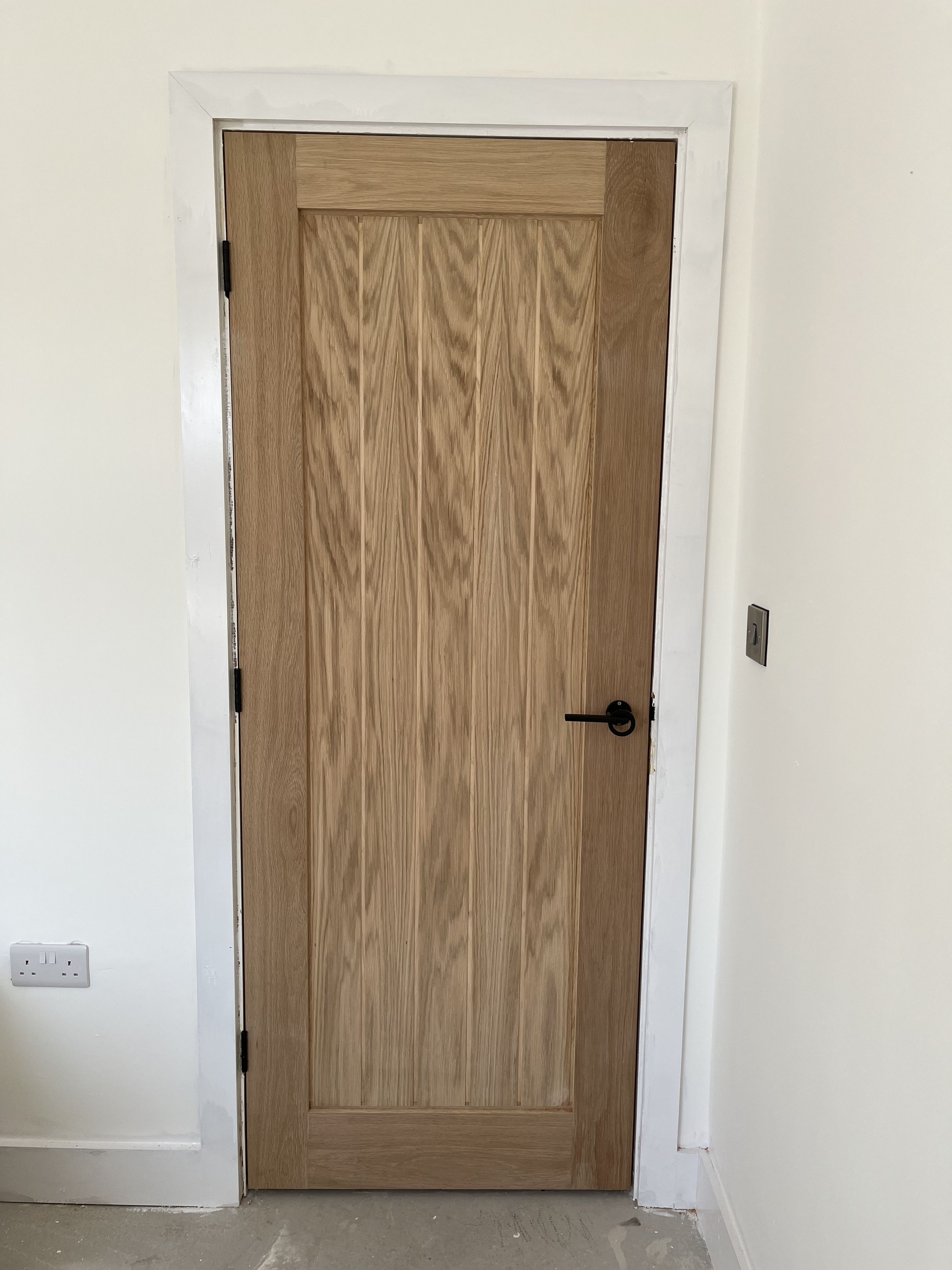 Wooden interior door with black handle and hinges, installed in white wall with electrical outlet on the left and switch on the right.