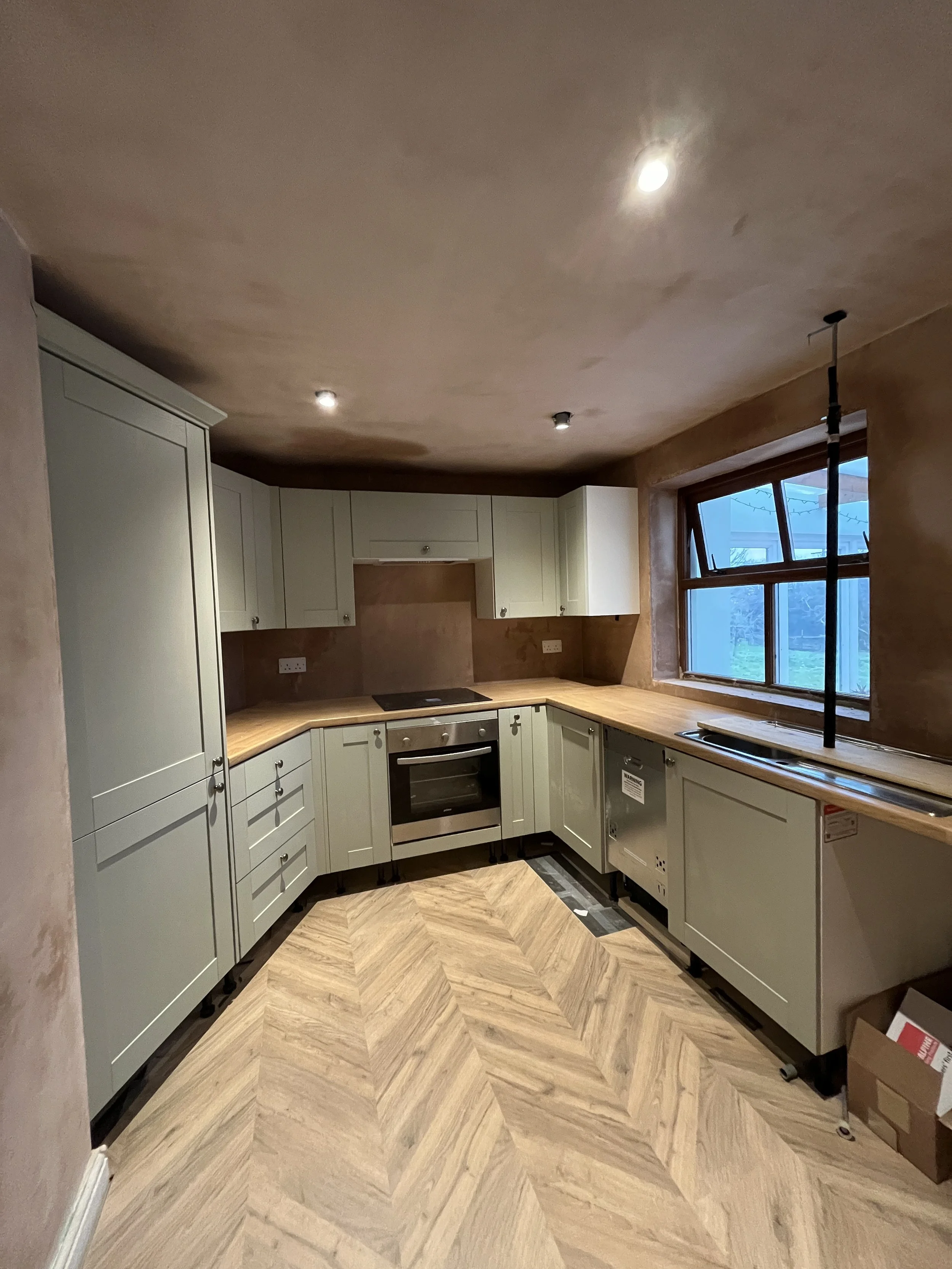 Kitchen with cream-colored cabinets, wood countertops, large window, and a space for appliances under construction. Recessed ceiling lights and a floor with a herringbone pattern.