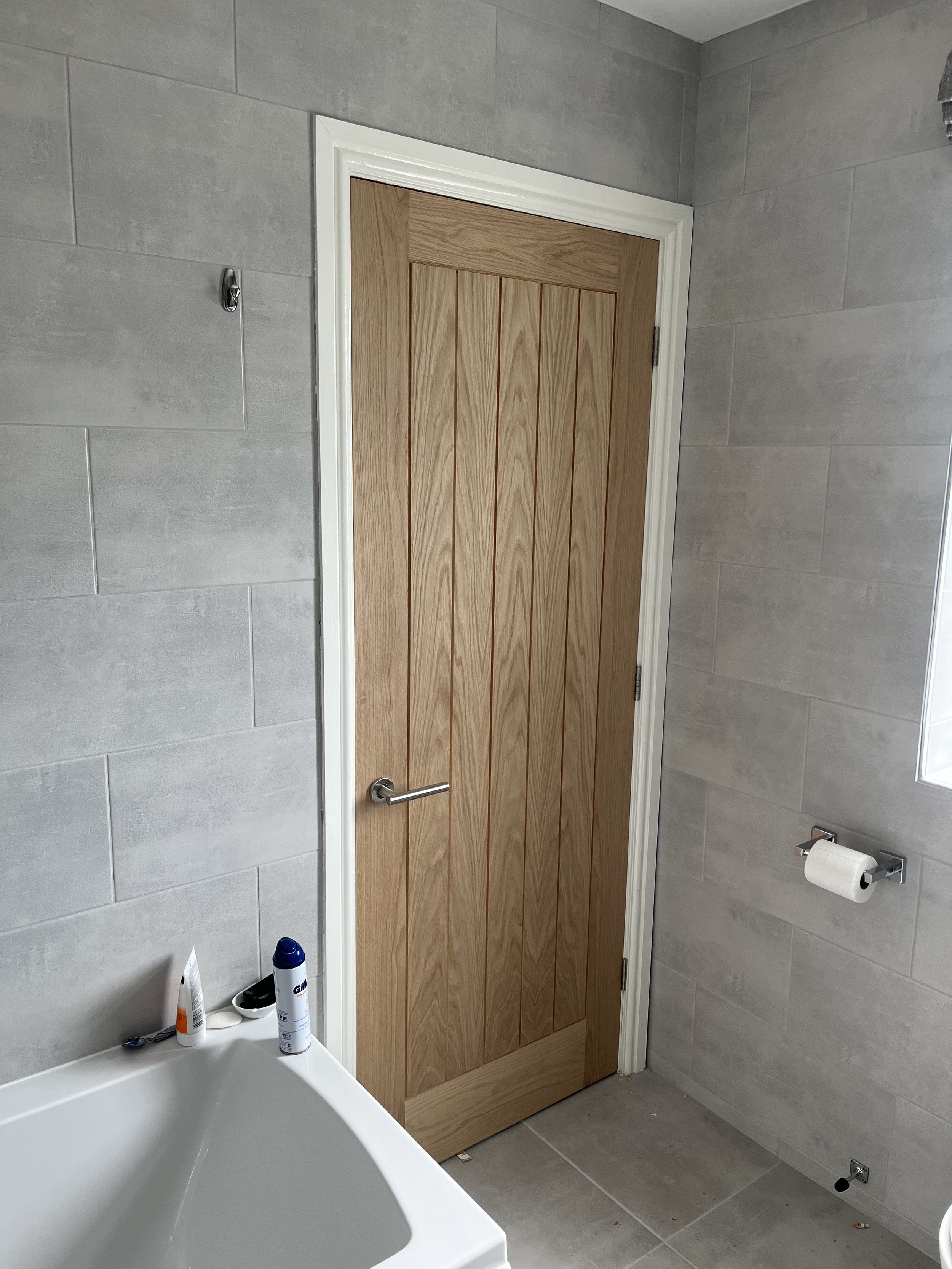 A bathroom with grey tiled walls, a wooden door with black handle, a toilet paper holder with roll, toiletries on a white surface, and a corner of a bathtub.