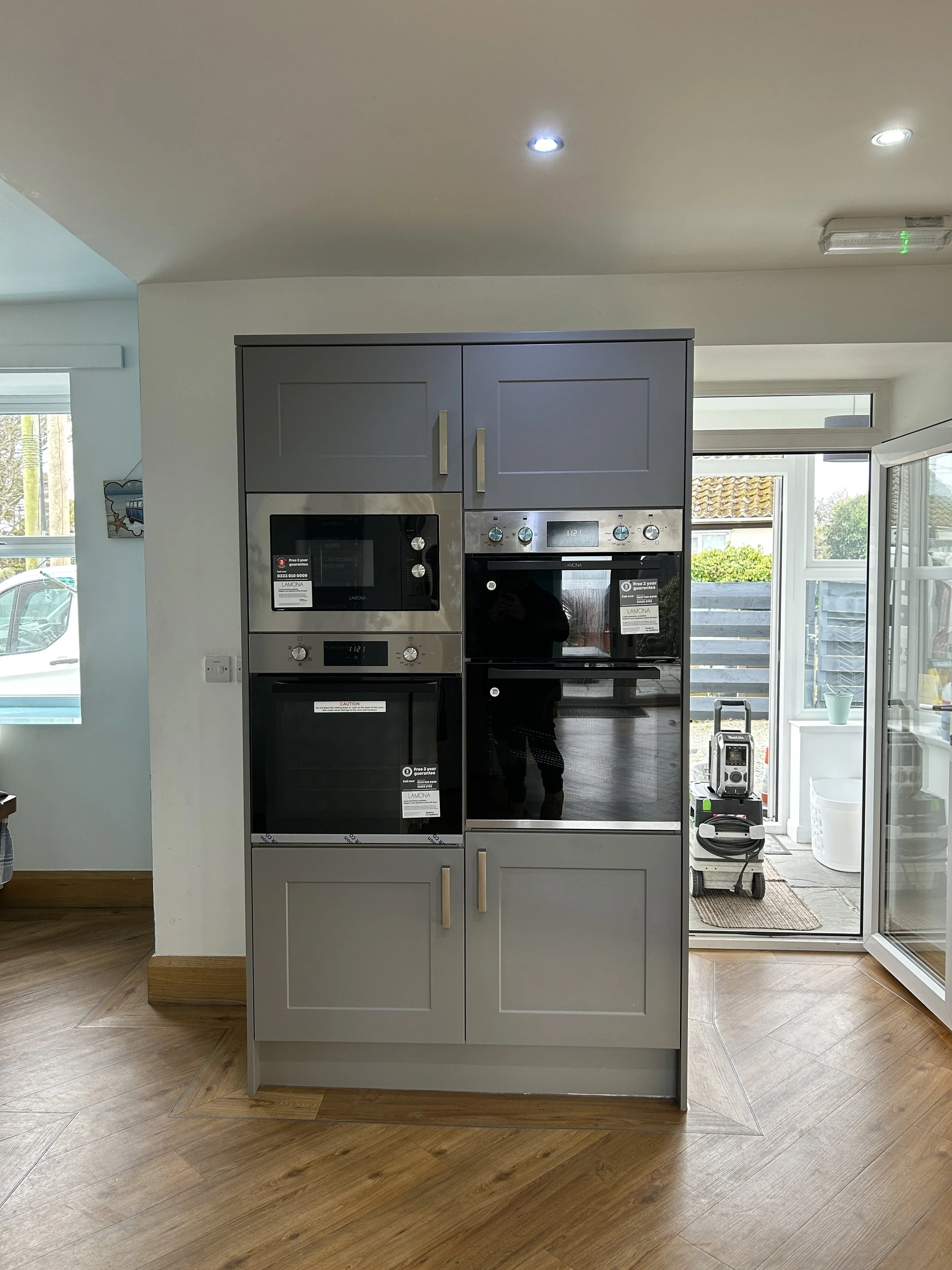 A kitchen with a grey cabinet housing an oven and microwave, hardwood floors, and a door to a patio with outdoor chairs and plants.