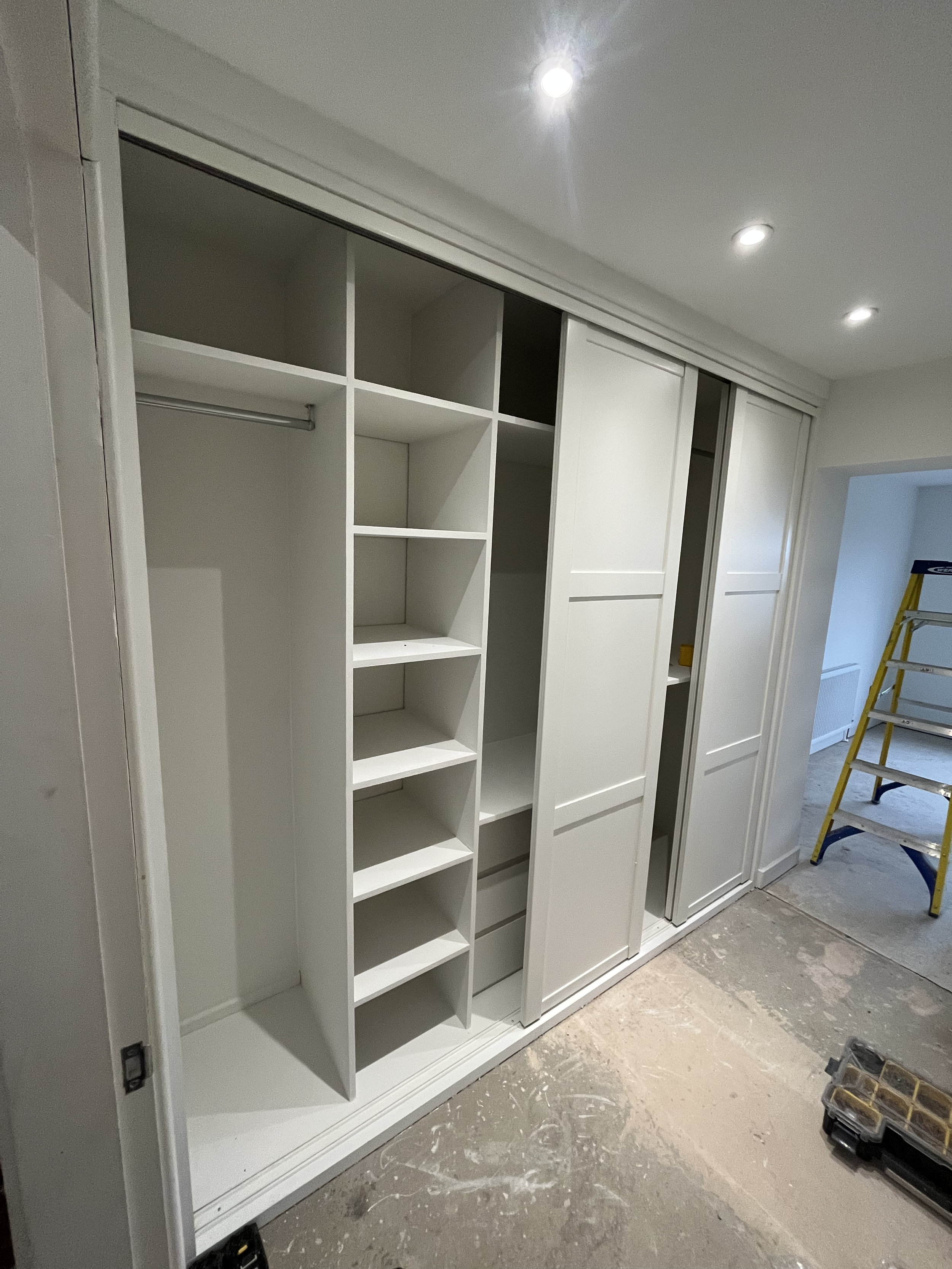 White built-in closet with open shelves, a hanging rod, and sliding doors, under construction, with a yellow ladder and work tools nearby.