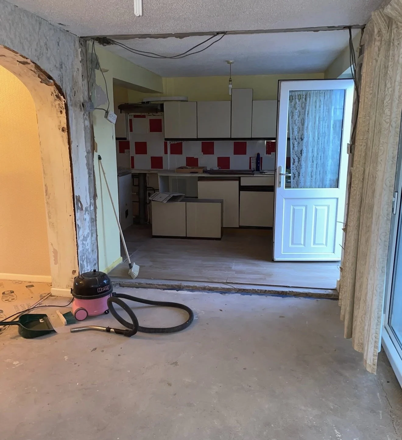 Room under renovation with a vacuum cleaner, vacuum hose, and cleaning tools on the floor, partially removed wall, and a view into the kitchen with cabinets, counter, and an open door leading outside.