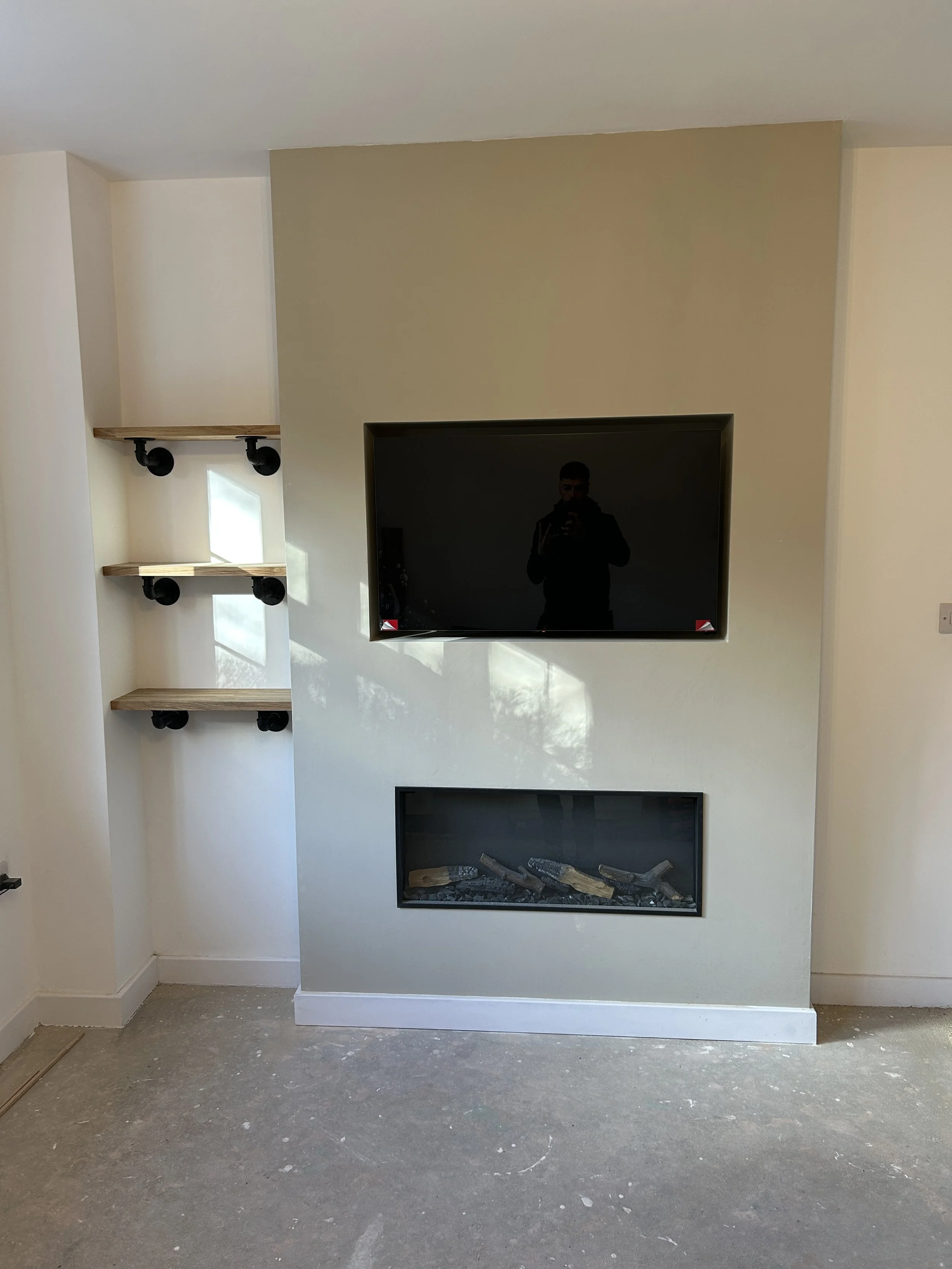 Modern living room wall with two wall-mounted shelves, a flat-screen TV, and a built-in fireplace at the bottom, with a reflection of a person taking the photo visible on the TV screen.