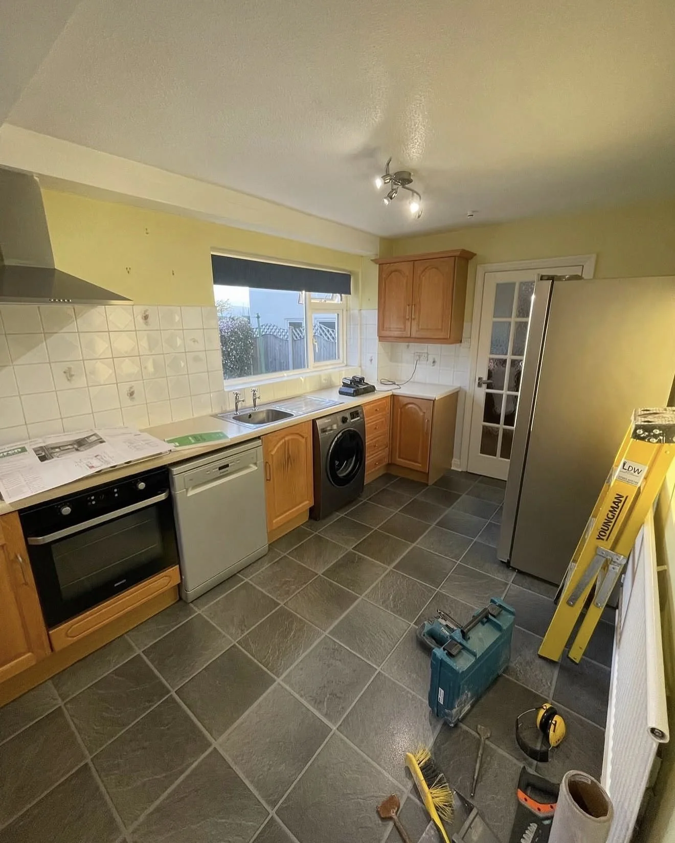 Kitchen with yellow walls, wooden cabinets, black and white appliances, tiled backsplash, window, door, and various tools and equipment on the floor.