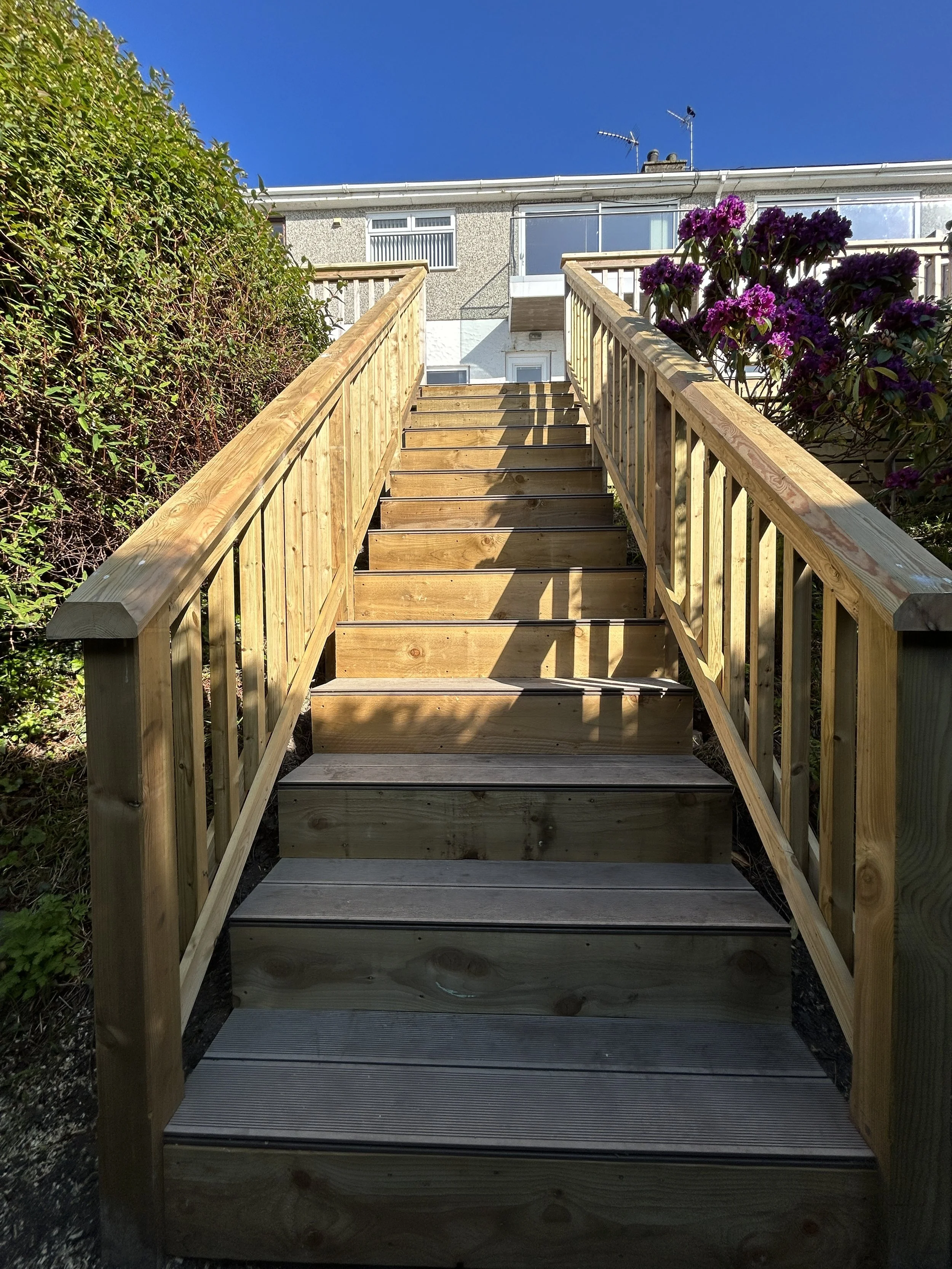 Wooden outdoor staircase with handrails leading up to a door on a residential building, with purple flowers on side and clear blue sky above.