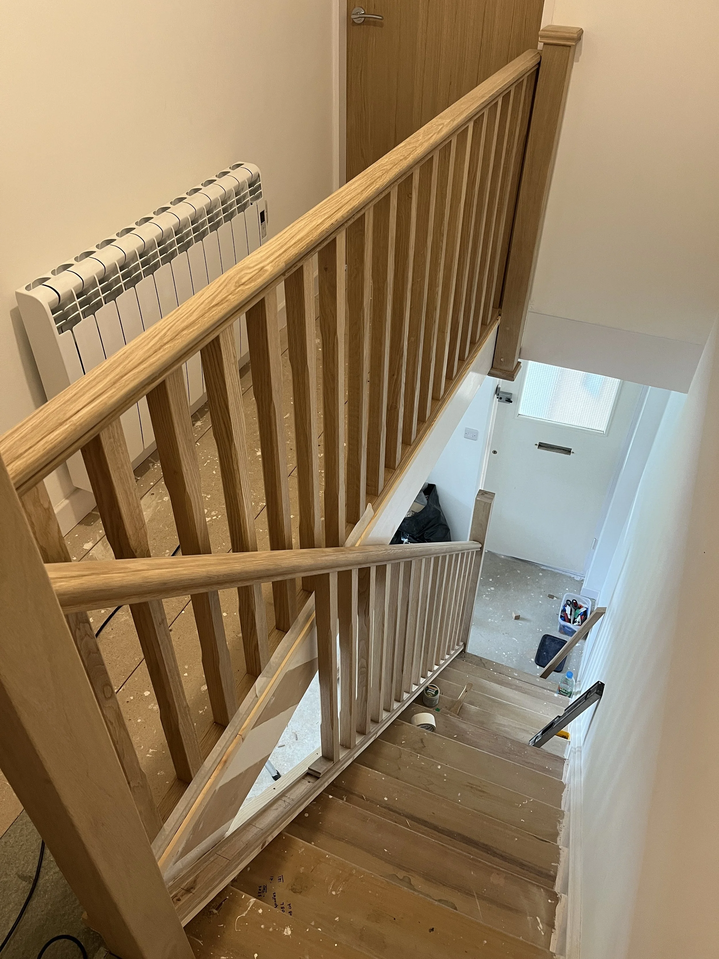 Wooden staircase with a landing, viewed from the top, with a staircase to the lower level visible at the bottom of the image; a white radiator on the wall to the left, and a door at the top of the staircase in the background.