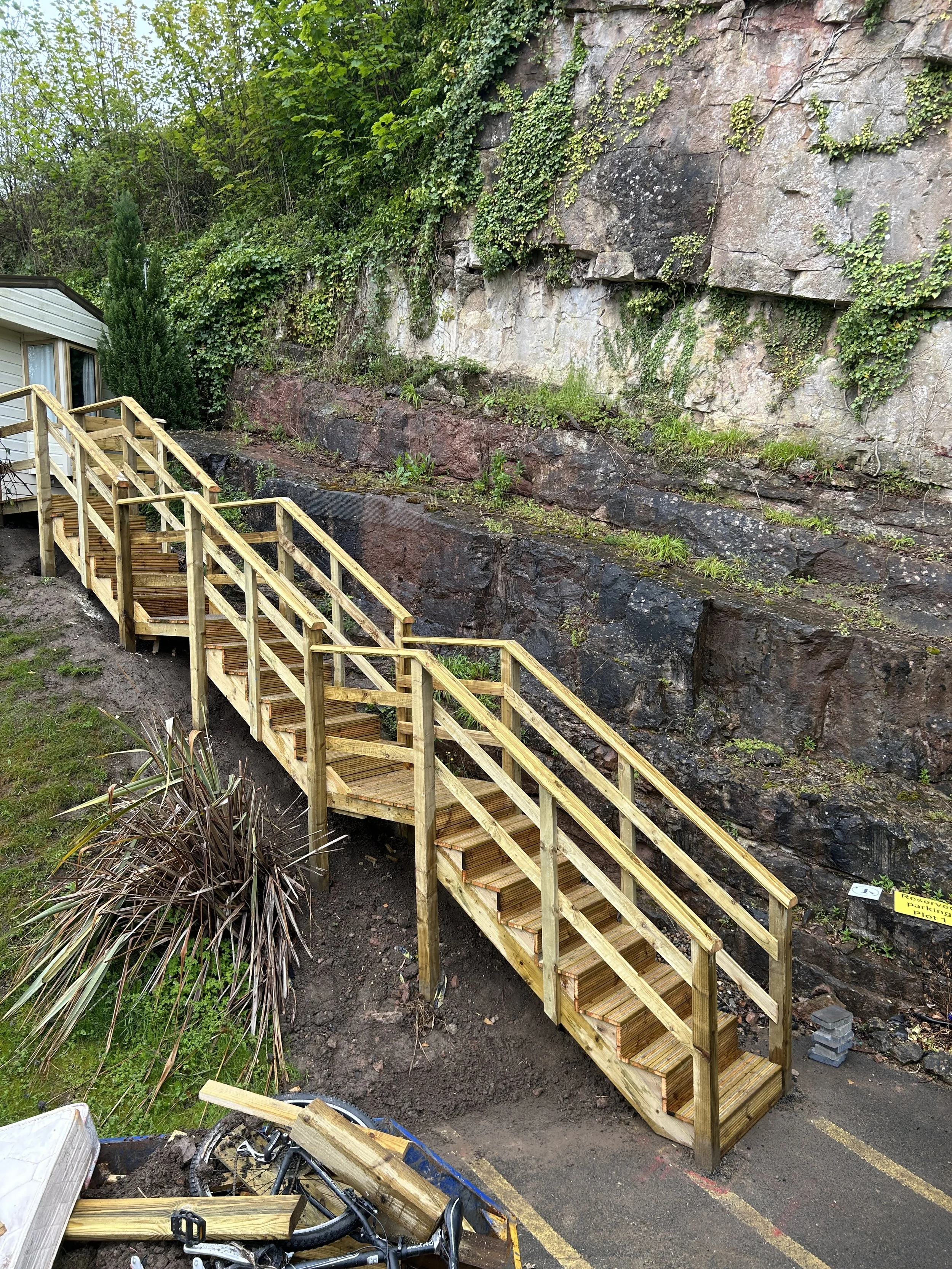 Wooden staircase with railing ascending beside a large rocky cliff face covered in green moss and ivy, near a white building and bicycle.
